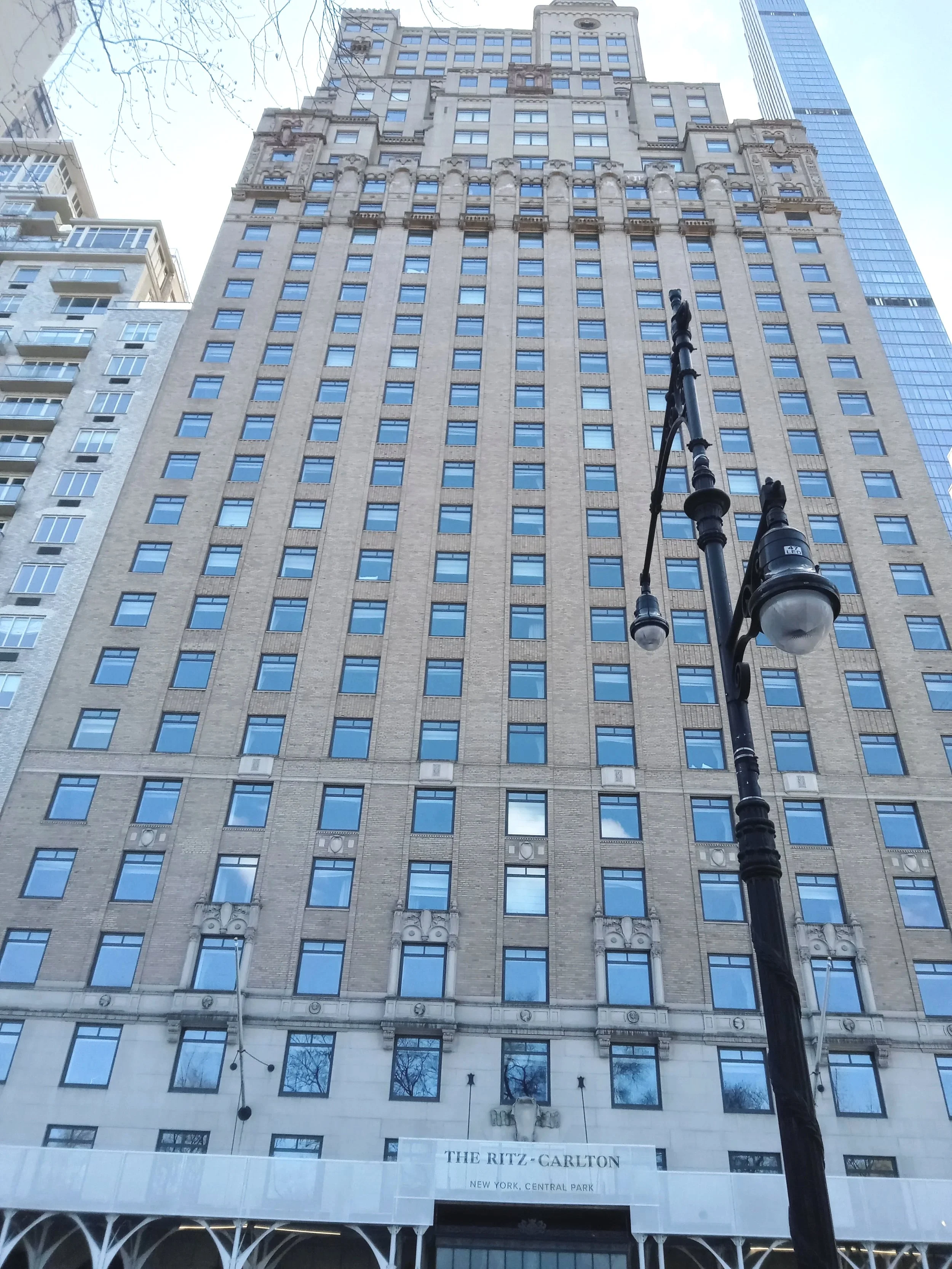 A tall historic building with numerous windows, a decorative facade, and a sign that reads 'The Ritz-Carlton, New York, Central Park' at the entrance, with a street lamp in the foreground and a modern skyscraper in the background.