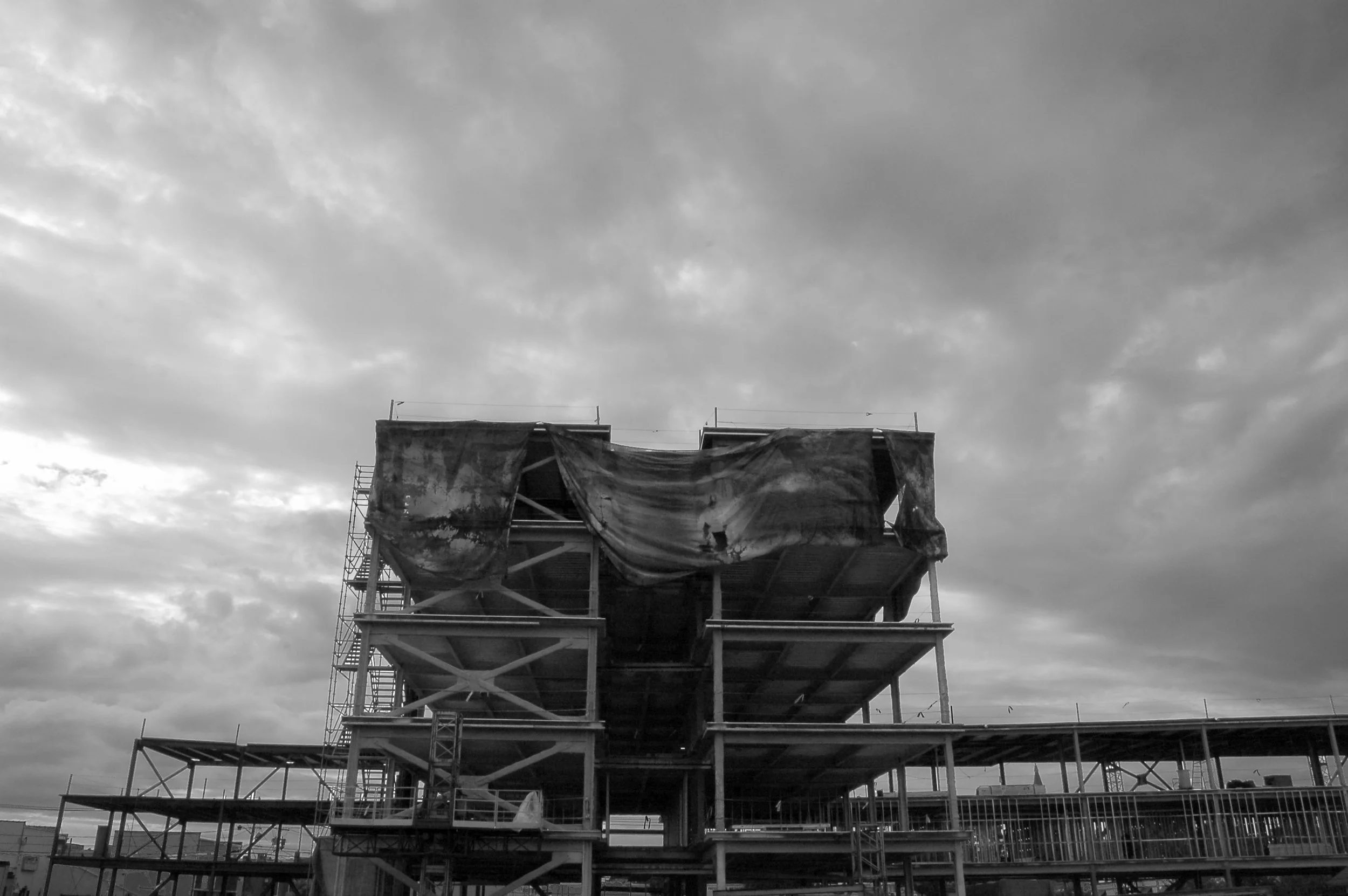 A multi-story building under construction with scaffolding and protective coverings, against a background of a cloudy sky.