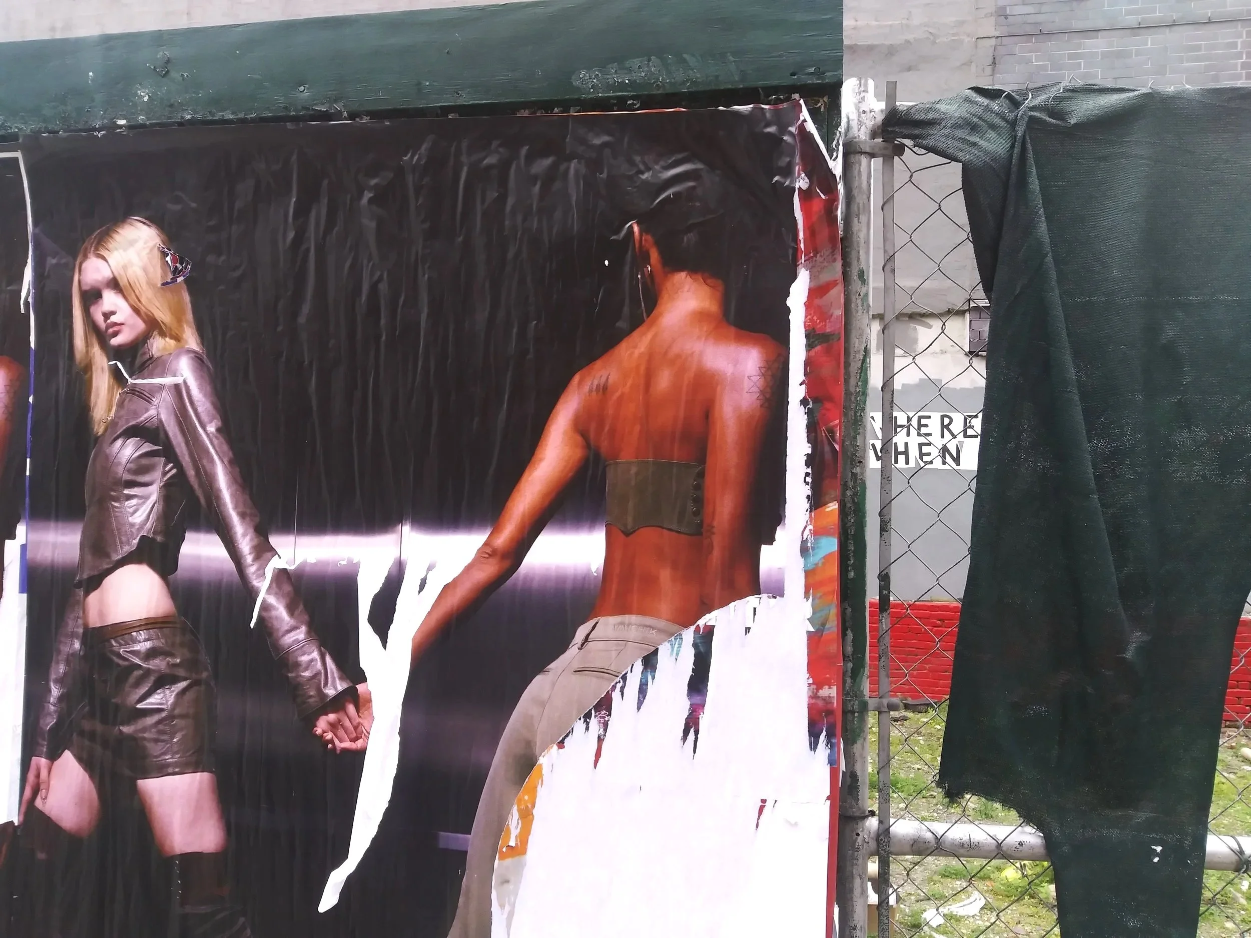 A torn poster featuring two models, a woman with long red hair wearing a leather jacket and shorts, and a topless woman with tattoos, at an outdoor urban setting.