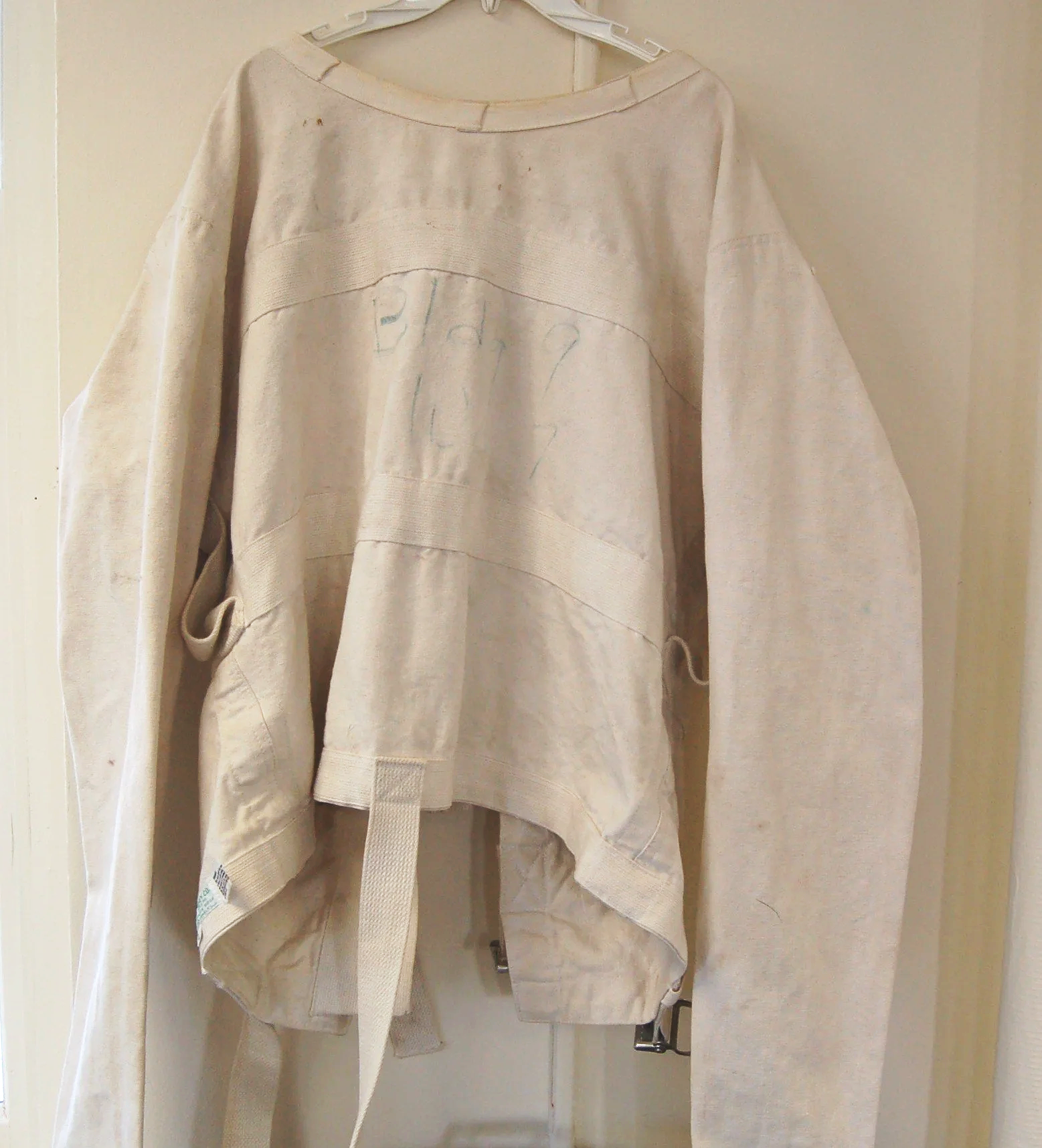 A beige or off-white oversized sweatshirt hanging on a white hanger against a beige wall. The sweatshirt appears to be made of a thick material with a round neckline and visible stitching details. There is faint writing or markings on the front, and a thick cloth belt hanging from the bottom.