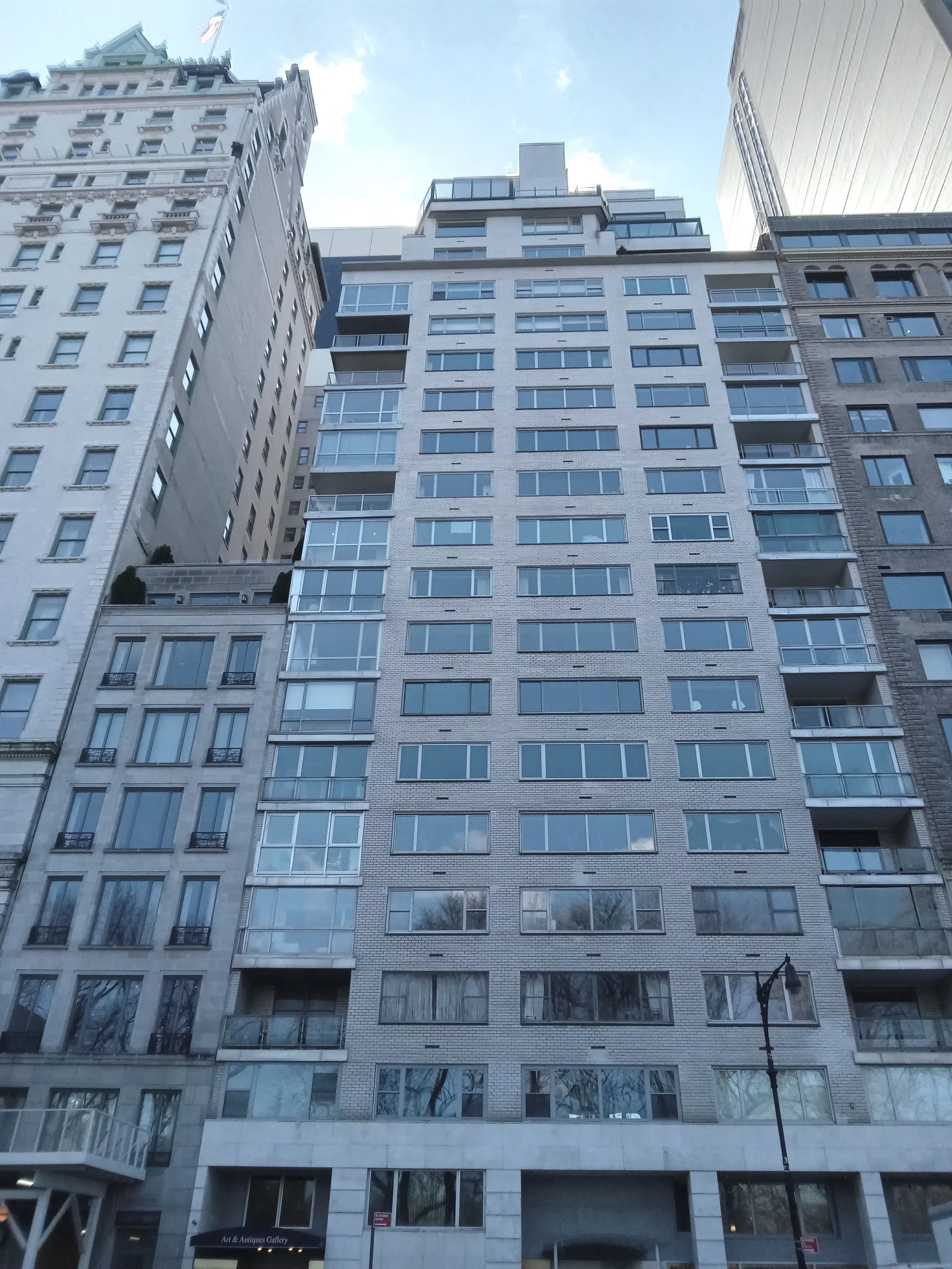 A tall city building with multiple floors, glass balconies, and a white brick facade, with neighboring buildings and a street lamp in front.