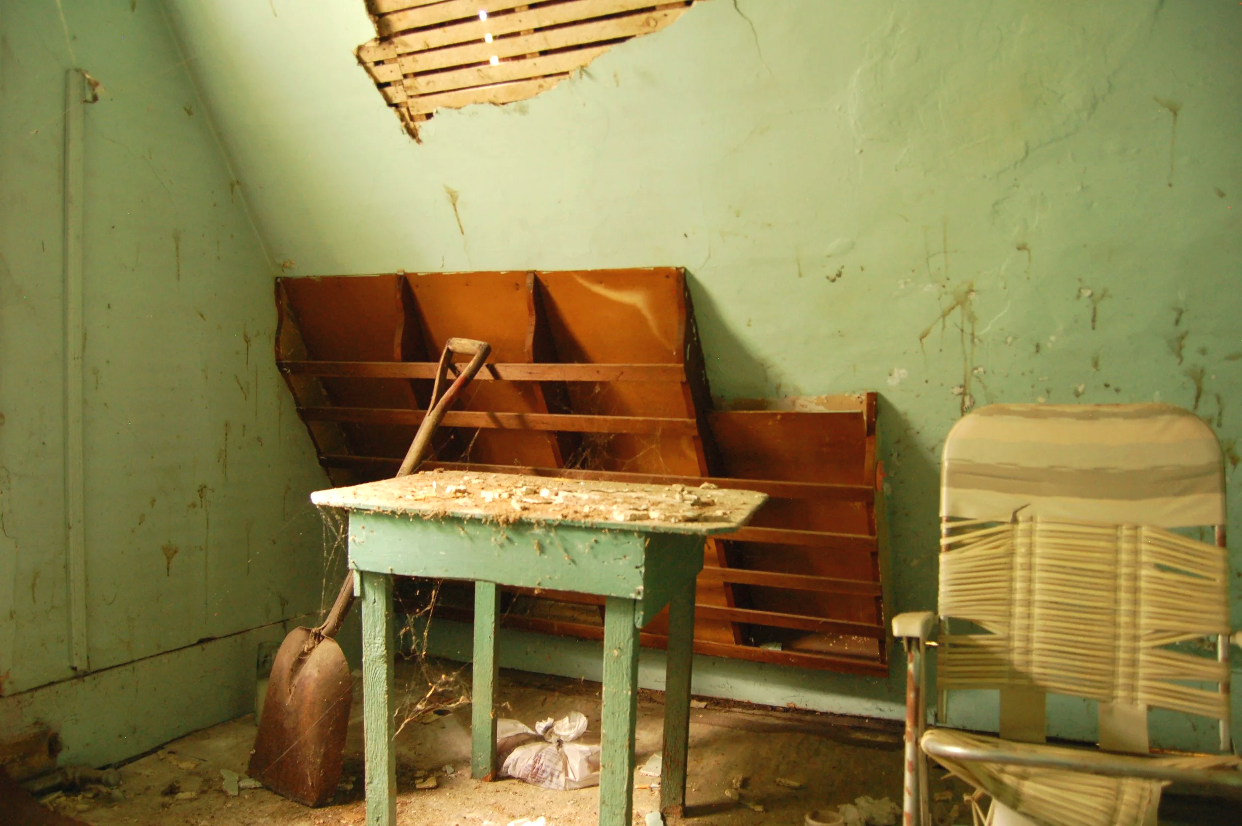 An abandoned room with peeling green paint on the walls, a battered green table with debris, an old bookshelf, and a stack of yellow plastic chairs, with a hole in the ceiling and cobwebs.