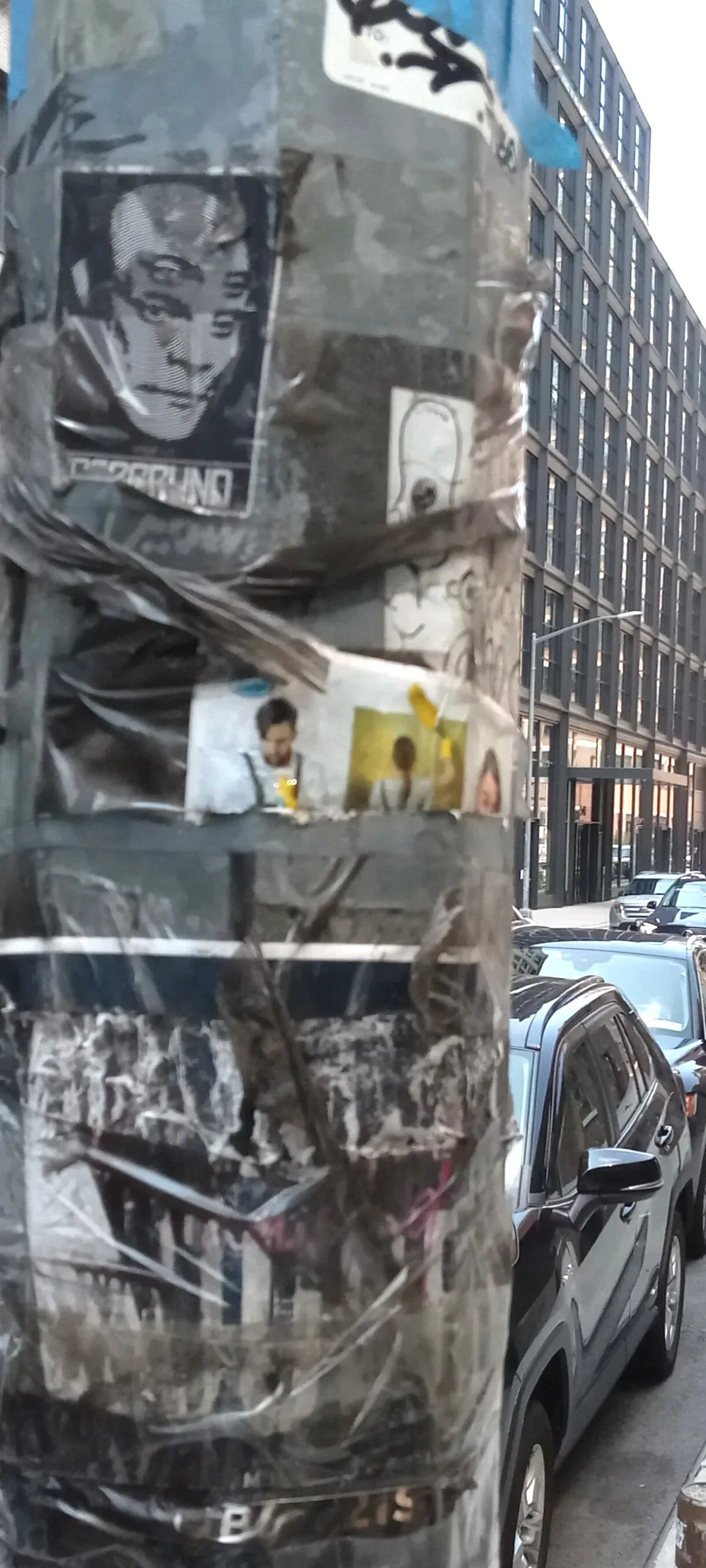 A close-up of a worn and damaged poster-covered pole on a city street with modern buildings and parked cars in the background.