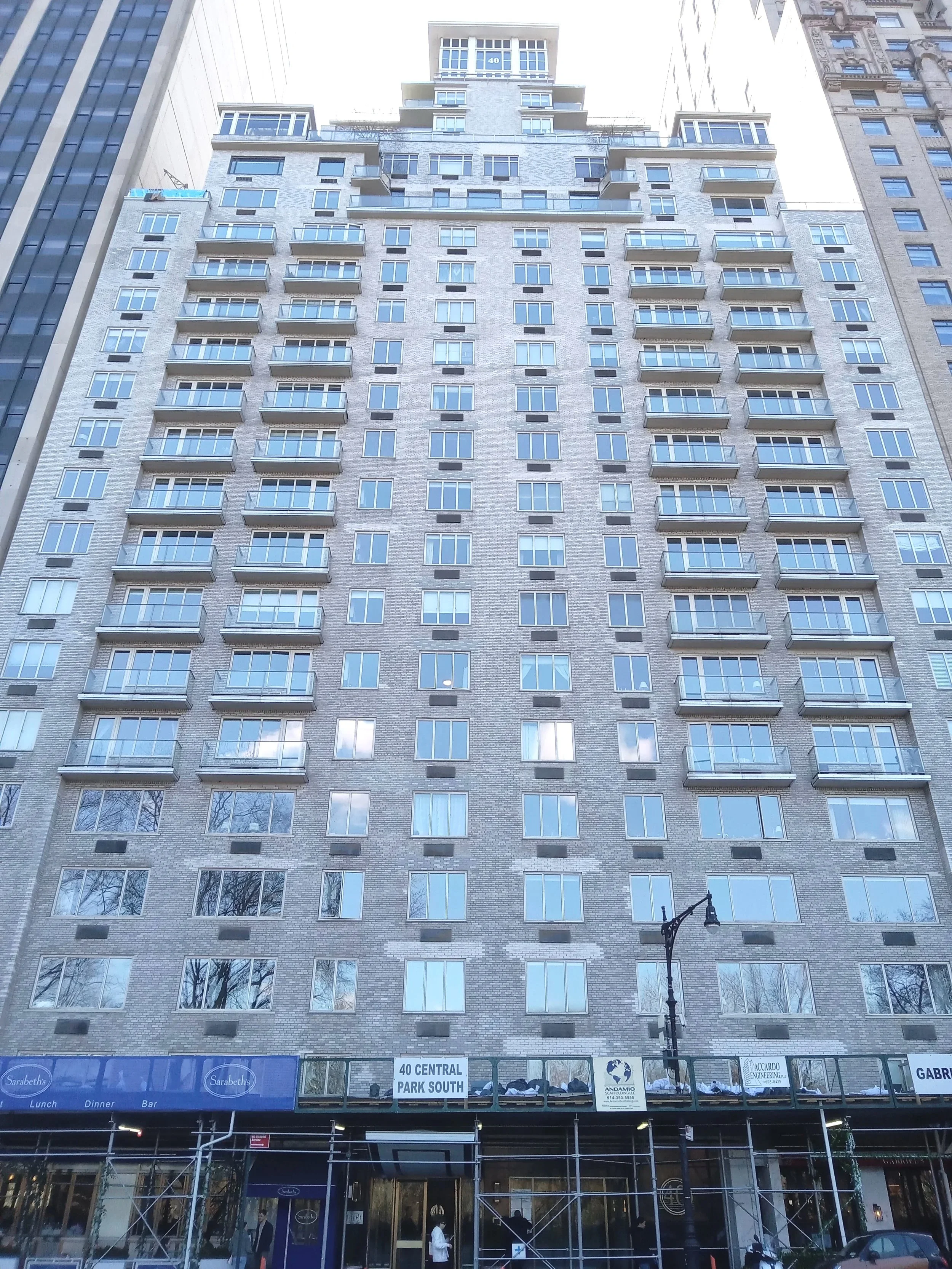 A tall multi-story building under construction with scaffolding at the bottom street level, signage indicating location as 40 Central, Park South, with a dark blue storefront named Sarabeth's and a street lamp.