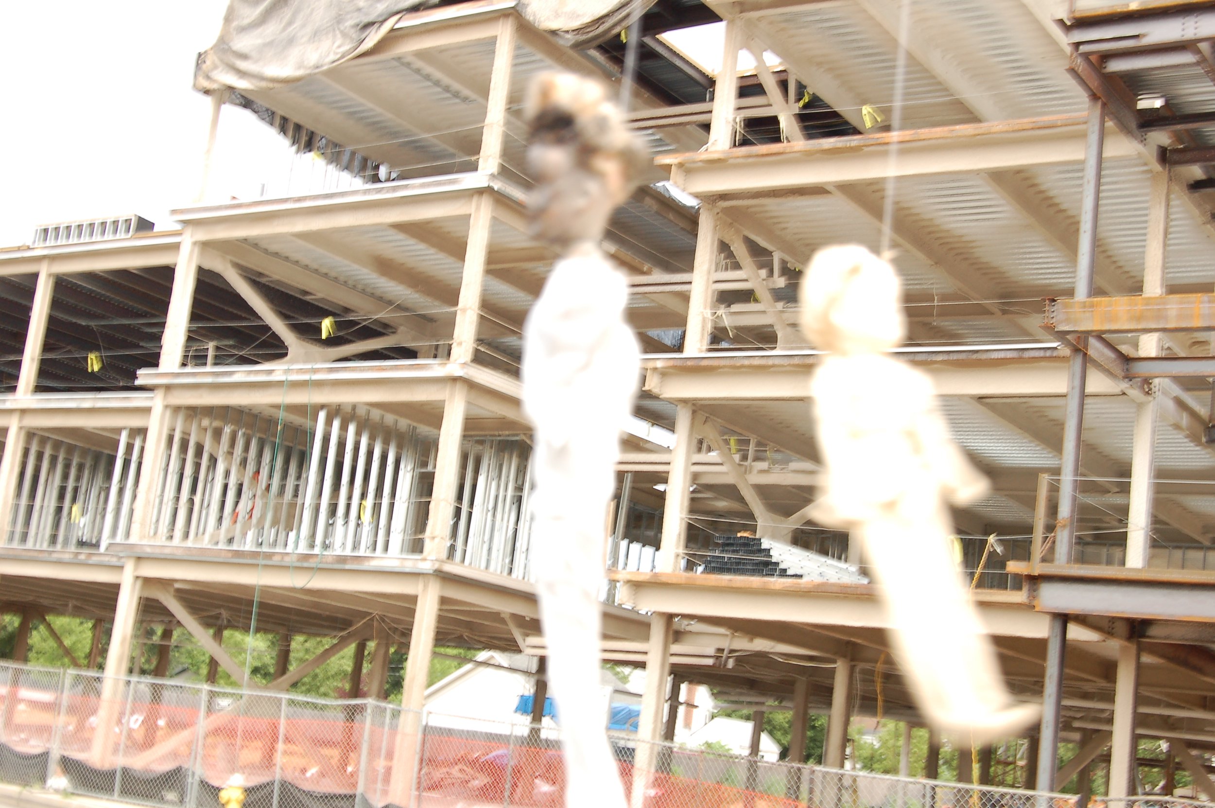 Construction site with unfinished building and hanging tan blocks, with a chain-link fence in the foreground and trees in the background.