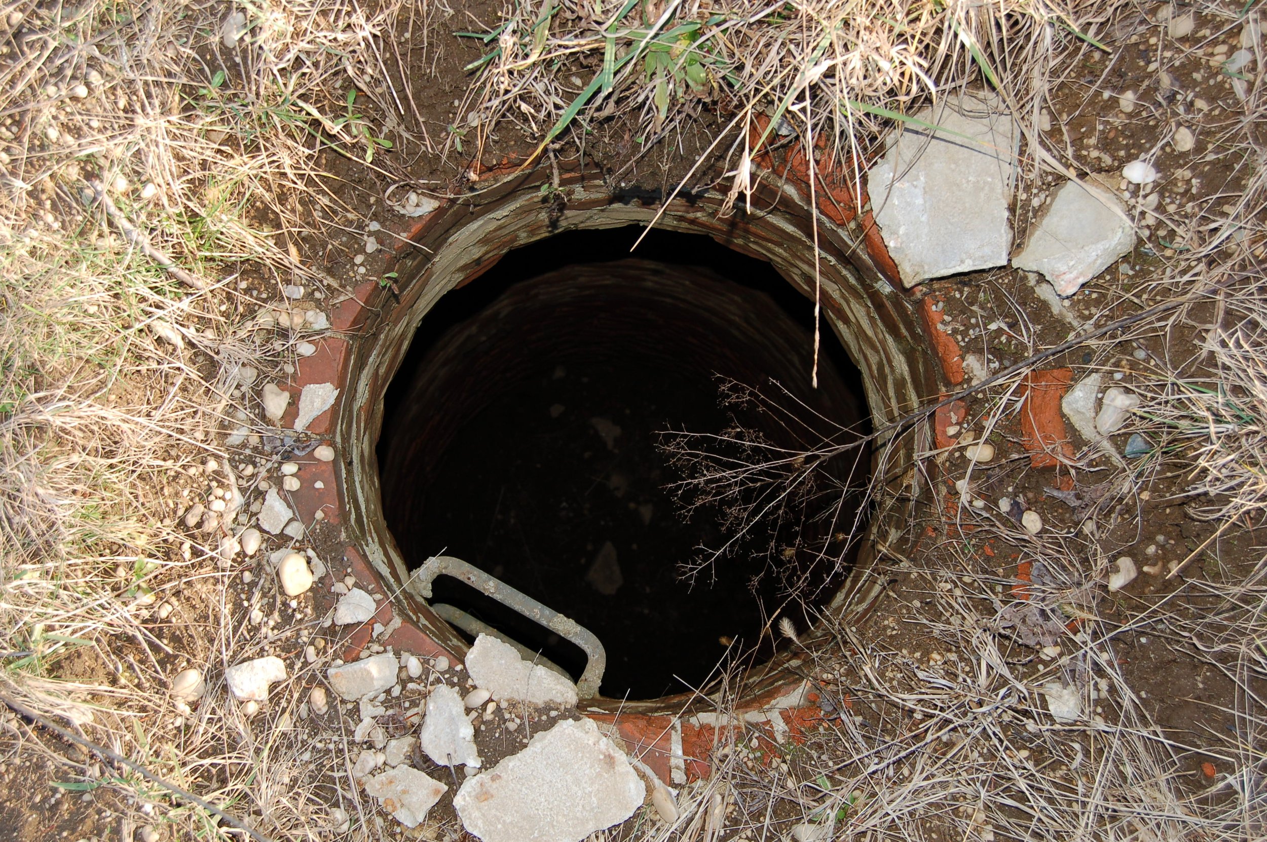 A hole with rocks and dirt around its opening, partially covered with dry grass and small stones.