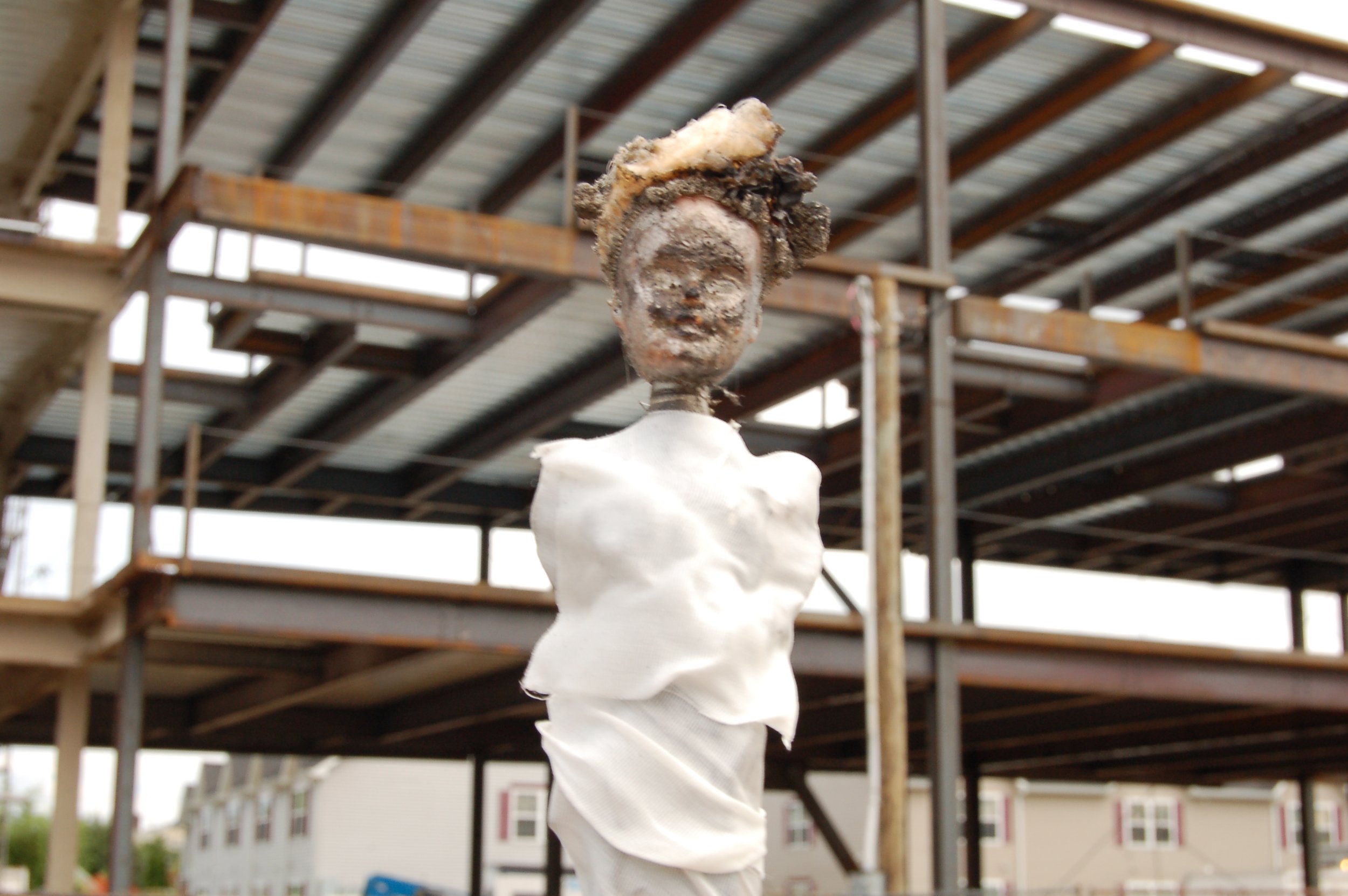 A statue of a woman's head with a large piece of material resembling hair or decoration on top, mounted on a white poseable material, set outdoors with a metal building structure in the background.