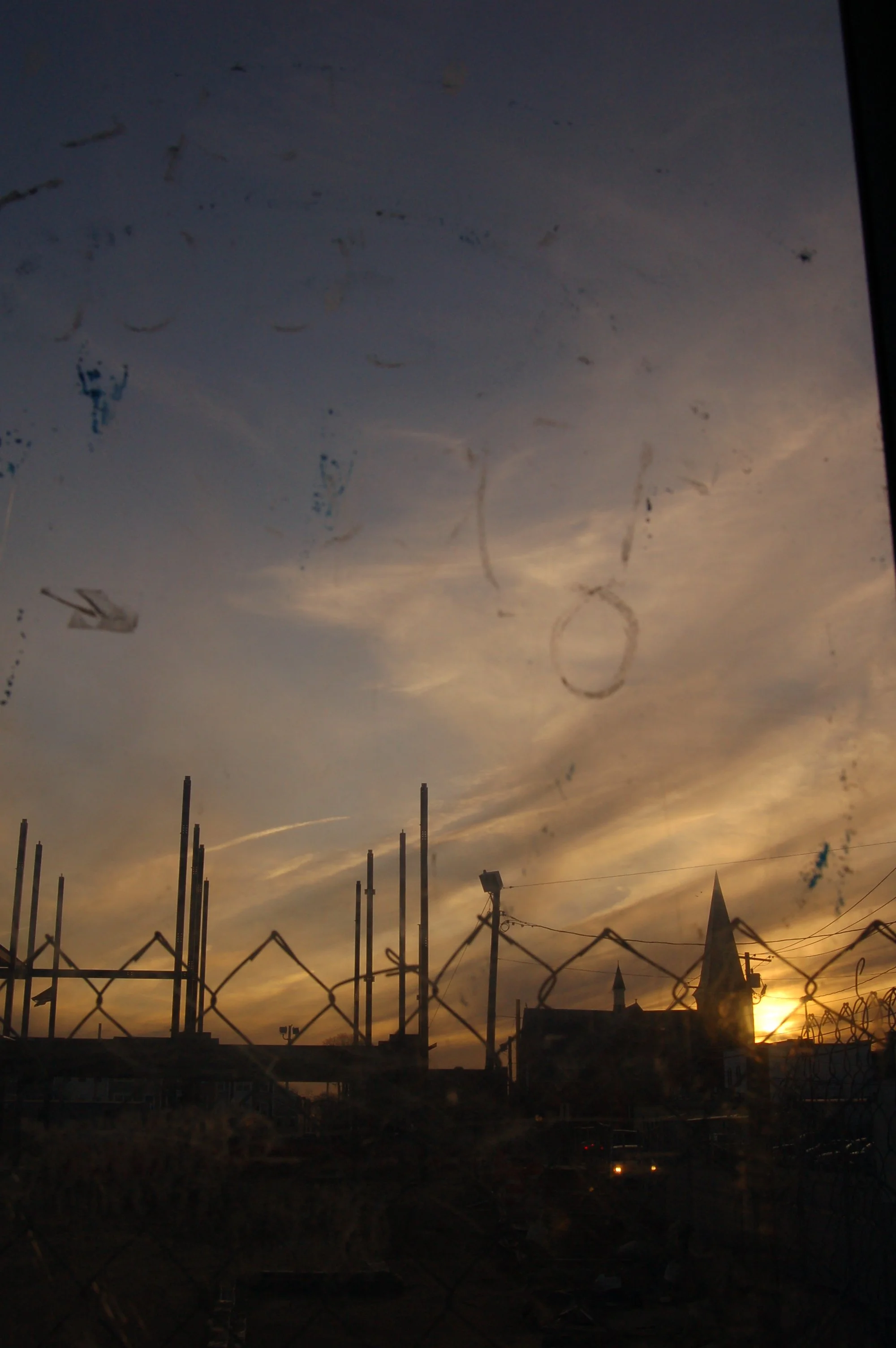 Sunset behind the skyline with church steeples, seen through a chain-link fence and a dirty window with streaks.