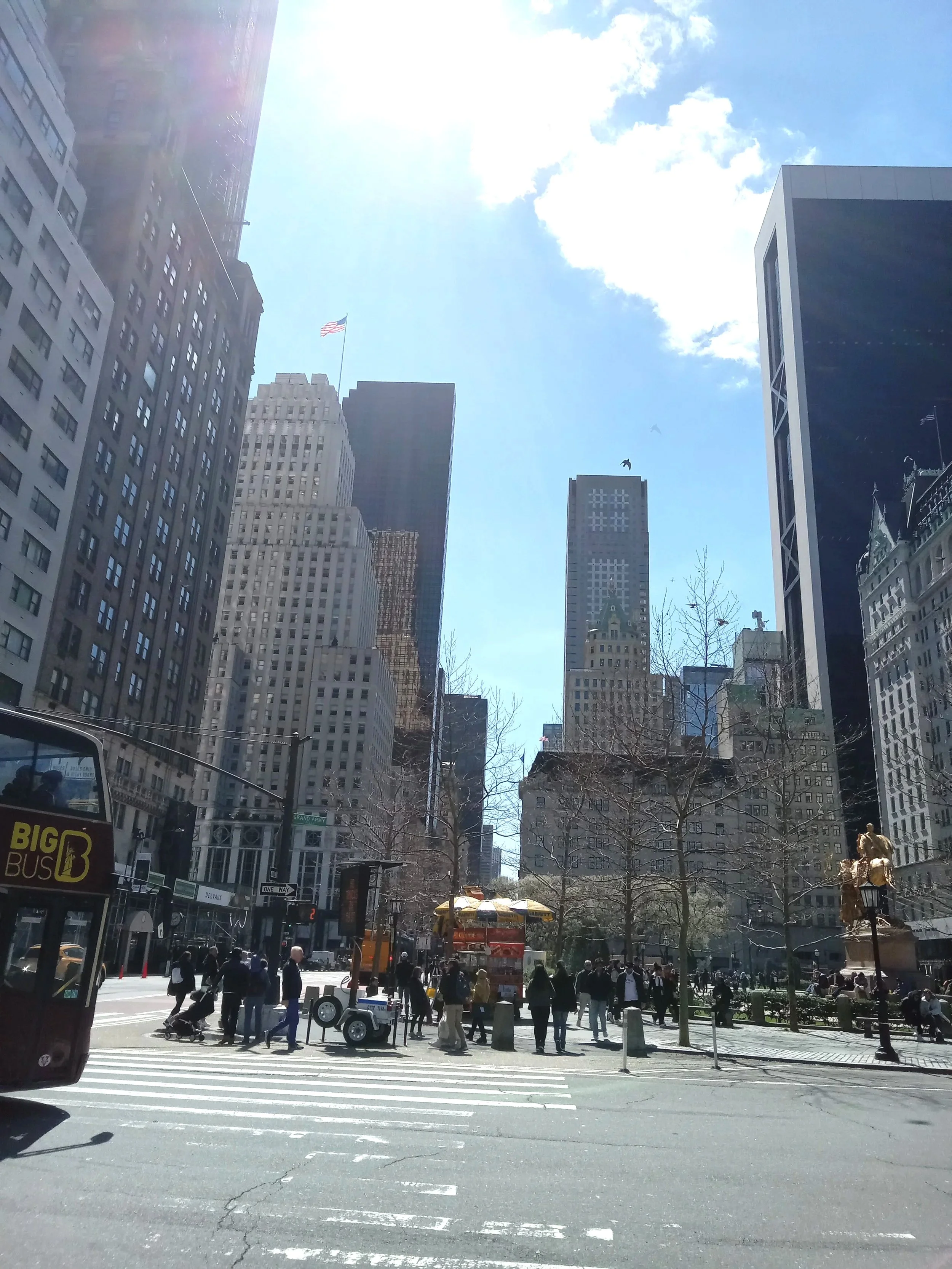 A busy city street scene with tall buildings, pedestrians crossing the street, and a yellow double-decker bus. An American flag is visible on one of the buildings under a bright blue sky with some clouds.