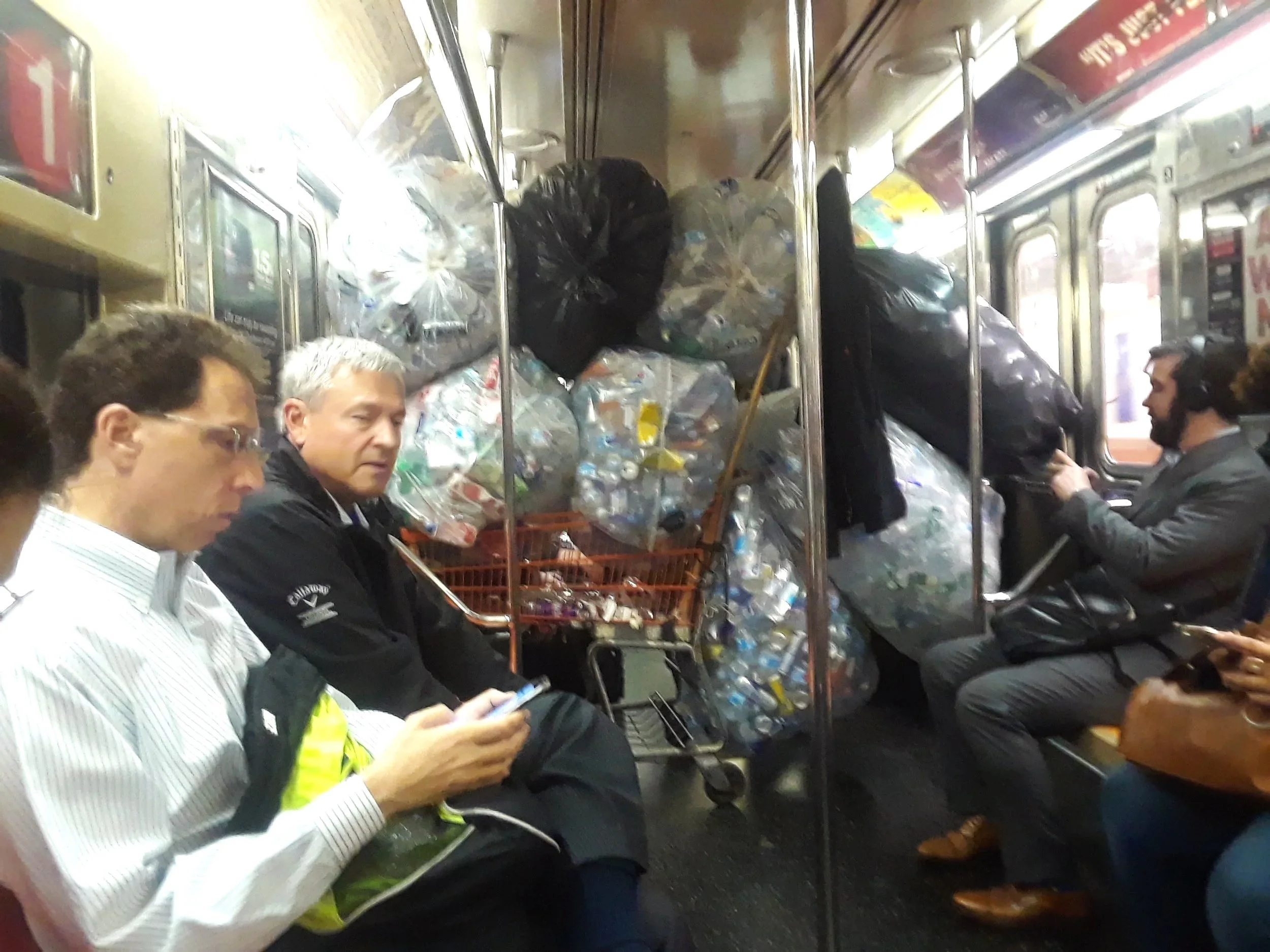 People sitting on a train, some using phones, with a pile of garbage bags of plastic bottles and recycling in the background.