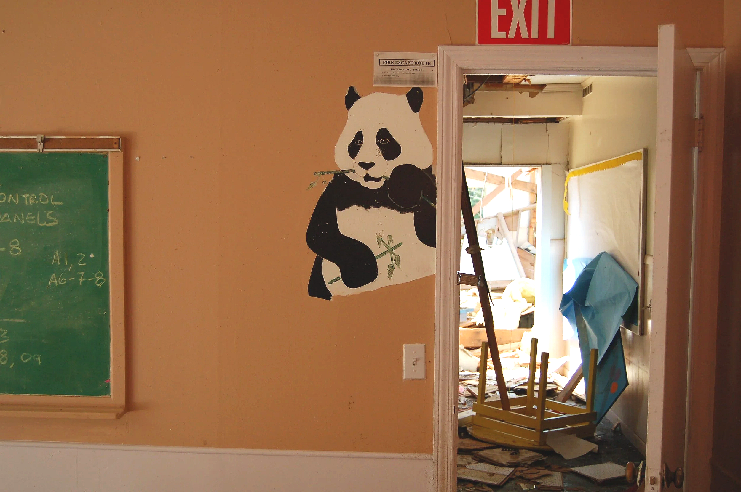 Wall with panda mural, doorway shows a construction site with debris and a framed fire escape route sign above the door. A green chalkboard is on the left side of the wall.