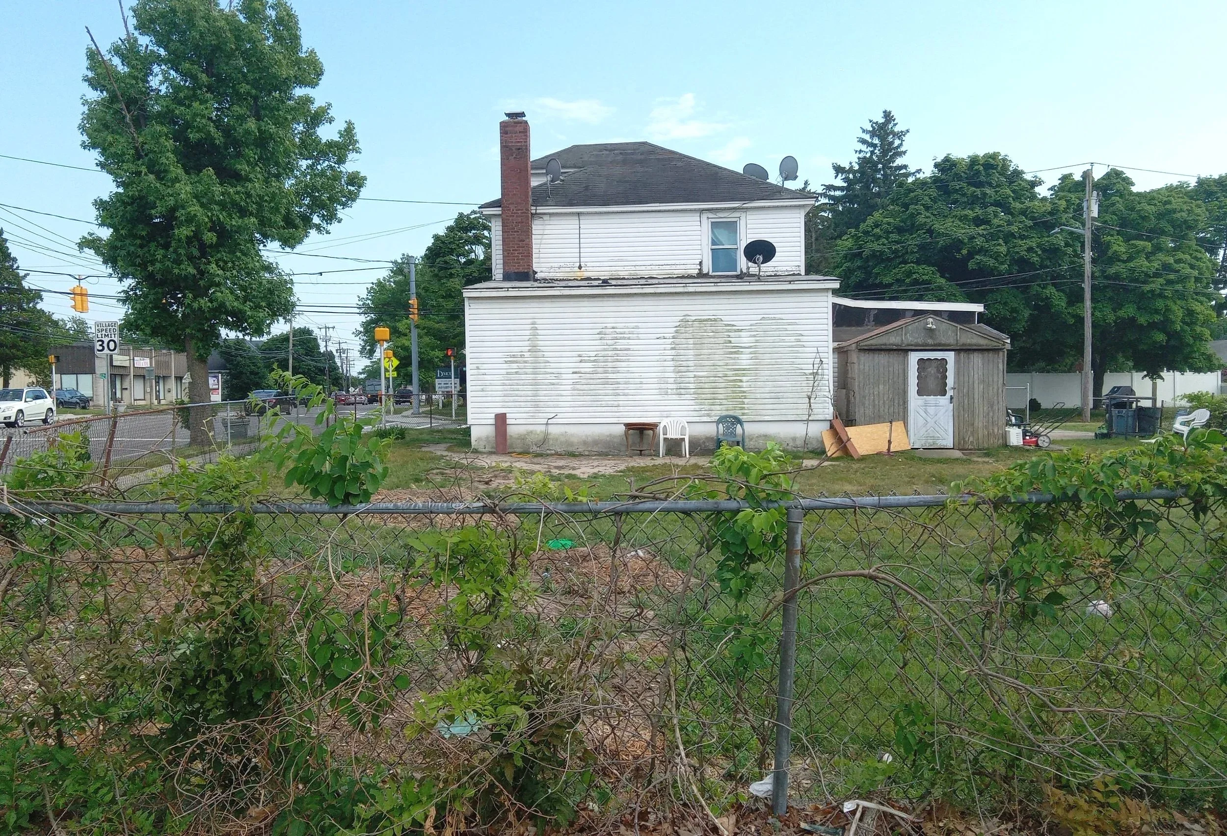 A two-story white house with a brick chimney, multiple satellite dishes on the roof, and peeling siding. In front, there is a small shed, outdoor chairs, and various yard items behind a chain-link fence. Trees and a busy street with cars and traffic lights are in the background.