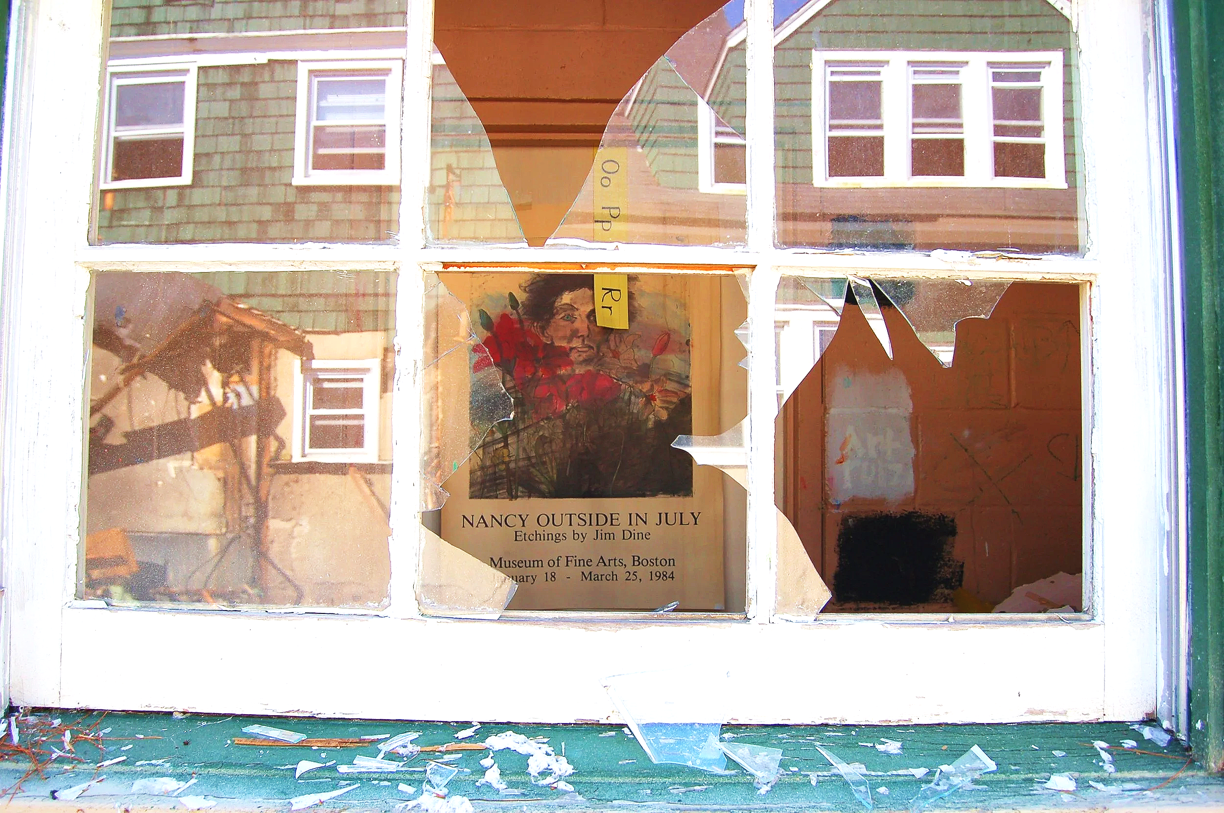 Broken window with shattered glass, a poster inside advertising an art exhibition titled 'Nancy Outside in July' at the Museum of Fine Arts, Boston, from January 18 to March 25, 1984.