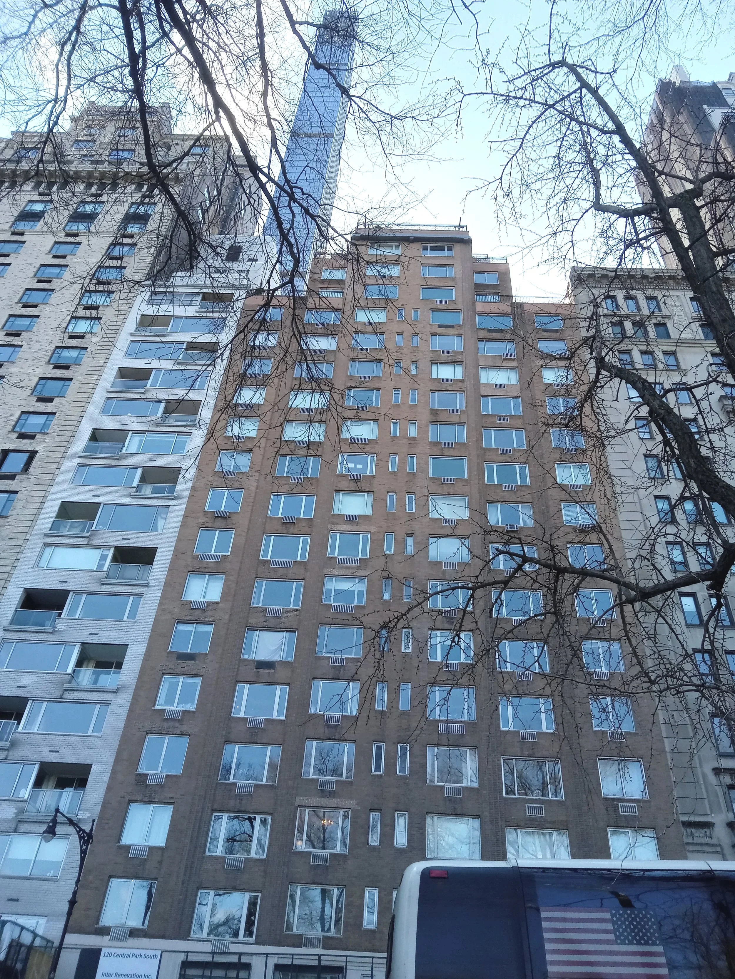 View of tall multi-story residential buildings with numerous windows, seen from street level, with leafless tree branches crossing the scene. A bus with an American flag sticker on the back is in the foreground.