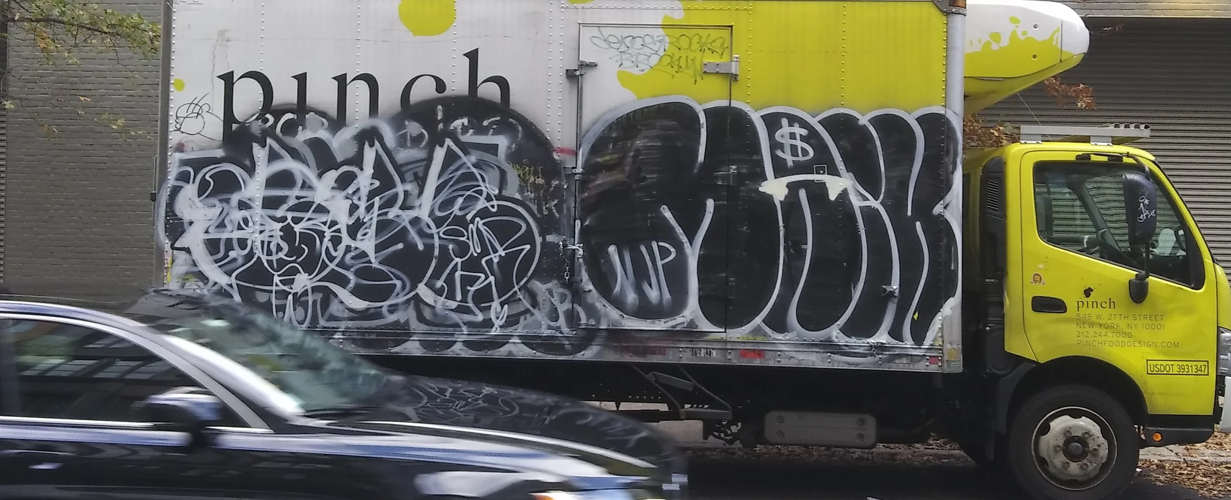 A yellow box truck with graffiti on its side, parked on the street. The graffitied side includes white and black tags, with the word 'punch' on the upper part and business information on the cab door.