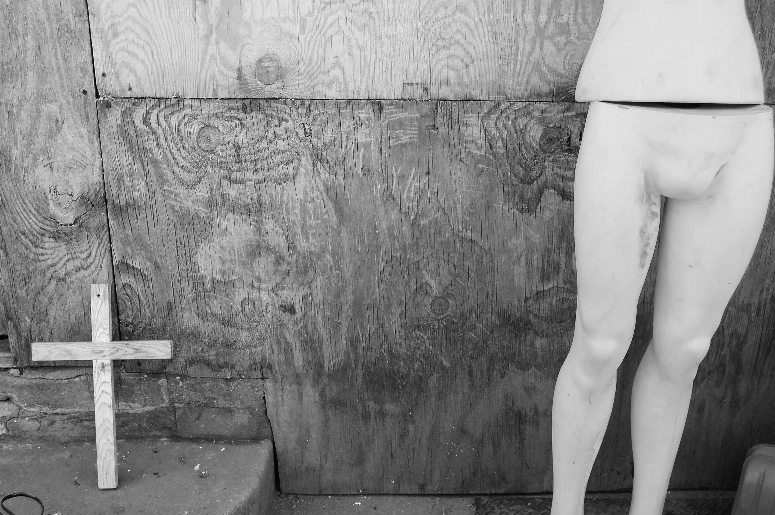 Part of a white mannequin with legs and hips visible, standing next to a wooden cross and an indistinct wooden background.