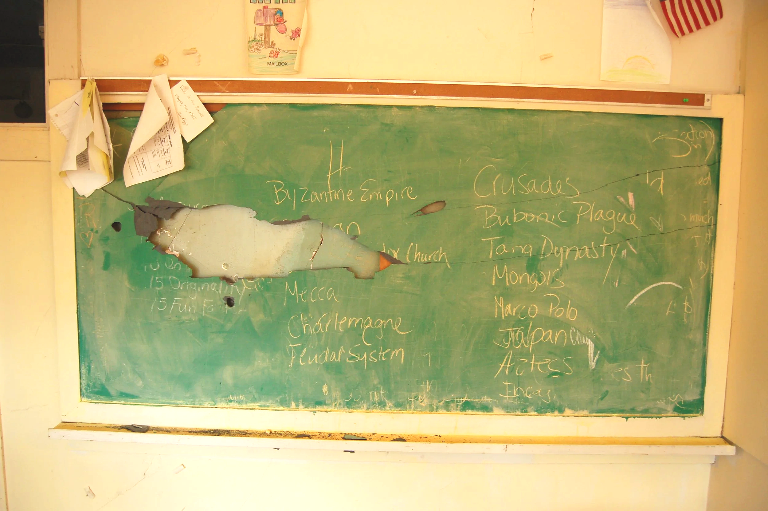 An old, damaged green chalkboard with chalk writings of historical empires and regions, partially obscured by torn paper, with a bulletin board above it and a small window on the side.