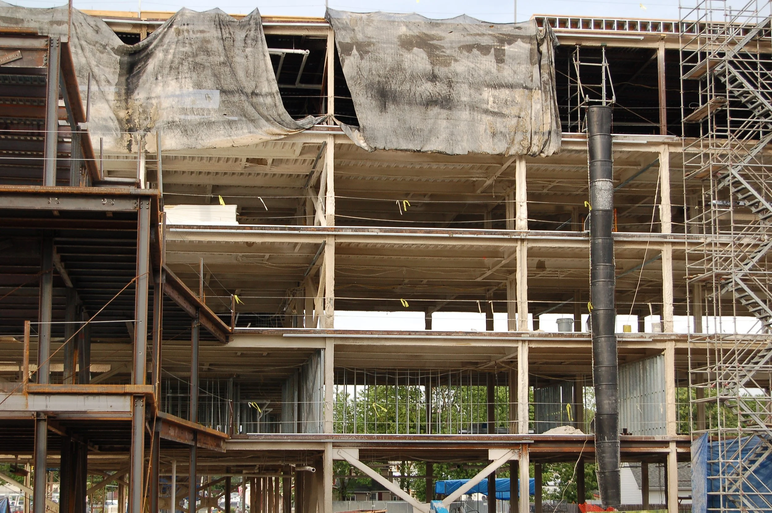 A multi-story building under construction with steel framing, scaffolding, and black plastic sheeting covering parts of the structure.