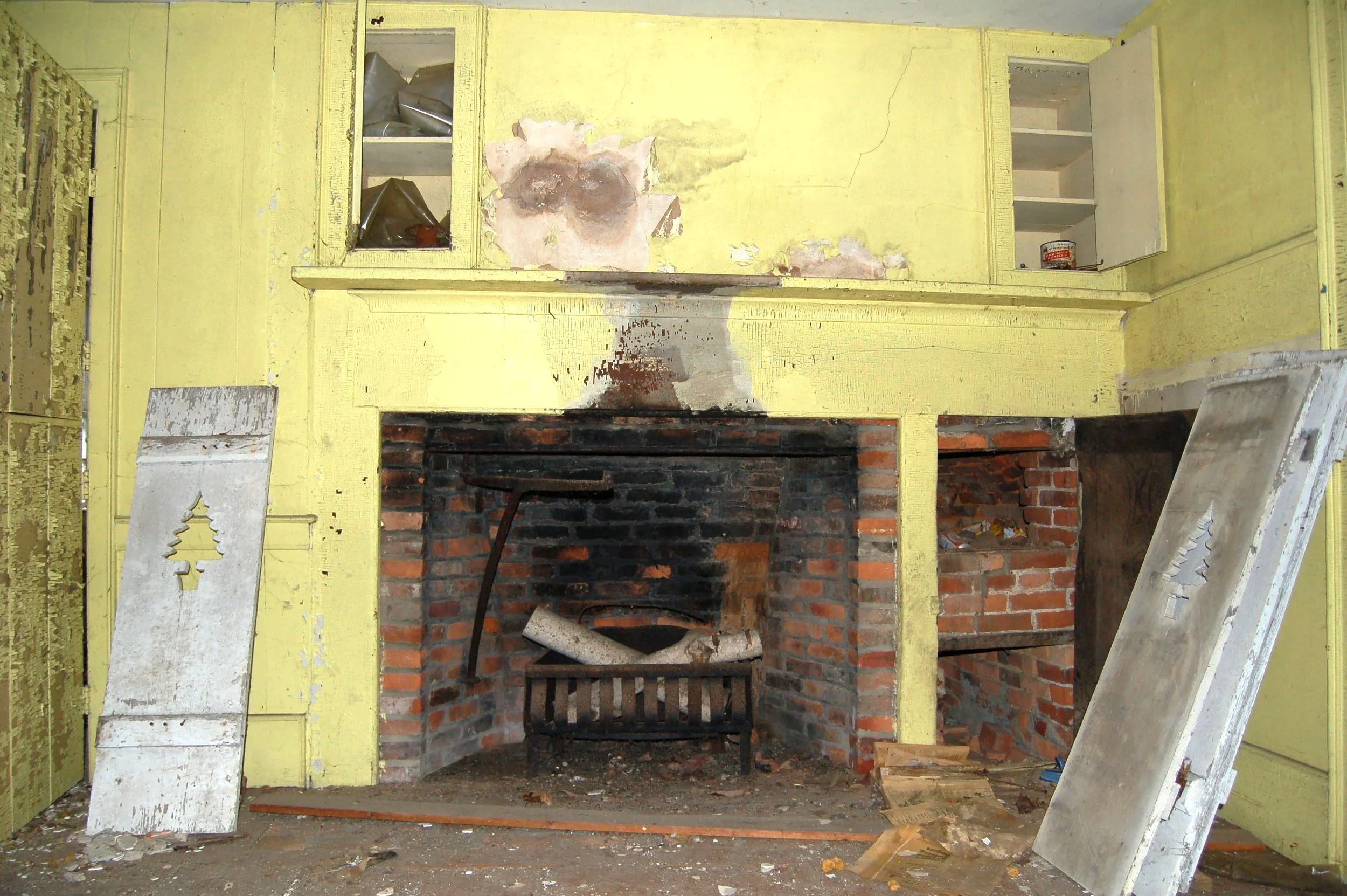 An underconstruction fireplace with yellow painted walls and open cabinets on each side, showing construction tools and materials, with a brick interior and debris on the floor.