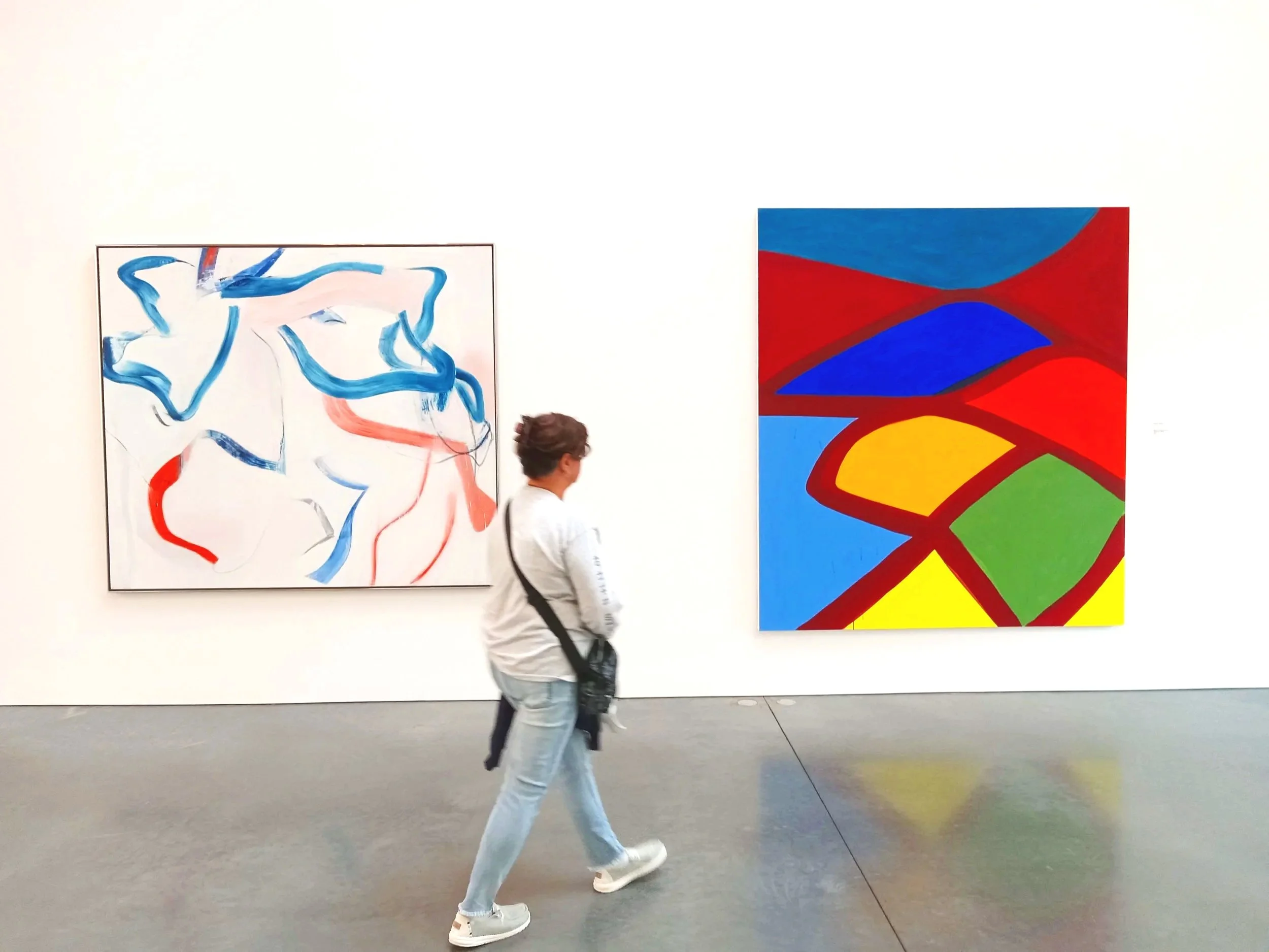 A person stands in front of abstract paintings in an art gallery. The paintings feature bold colors and shapes; one has blue and red outlines, and the other has geometric red, blue, green, yellow, and black areas.