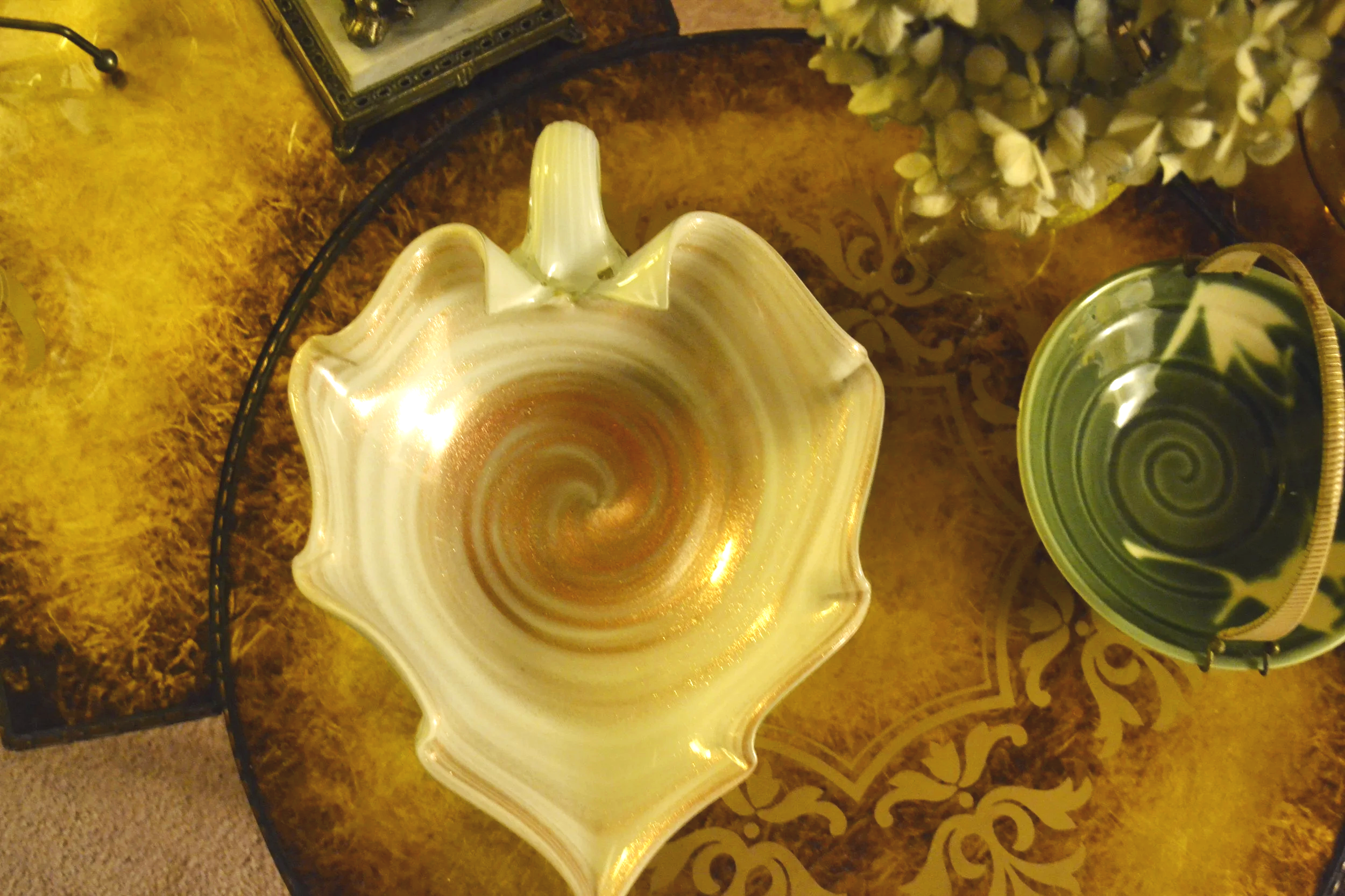 A decorative glass bowl with a gold-colored swirl pattern on a wooden table, surrounded by other decorative items including a green and beige ceramic piece.