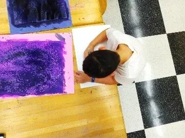 Person with short dark hair wearing a white shirt, seen from above, standing in front of a colorful textured painting.