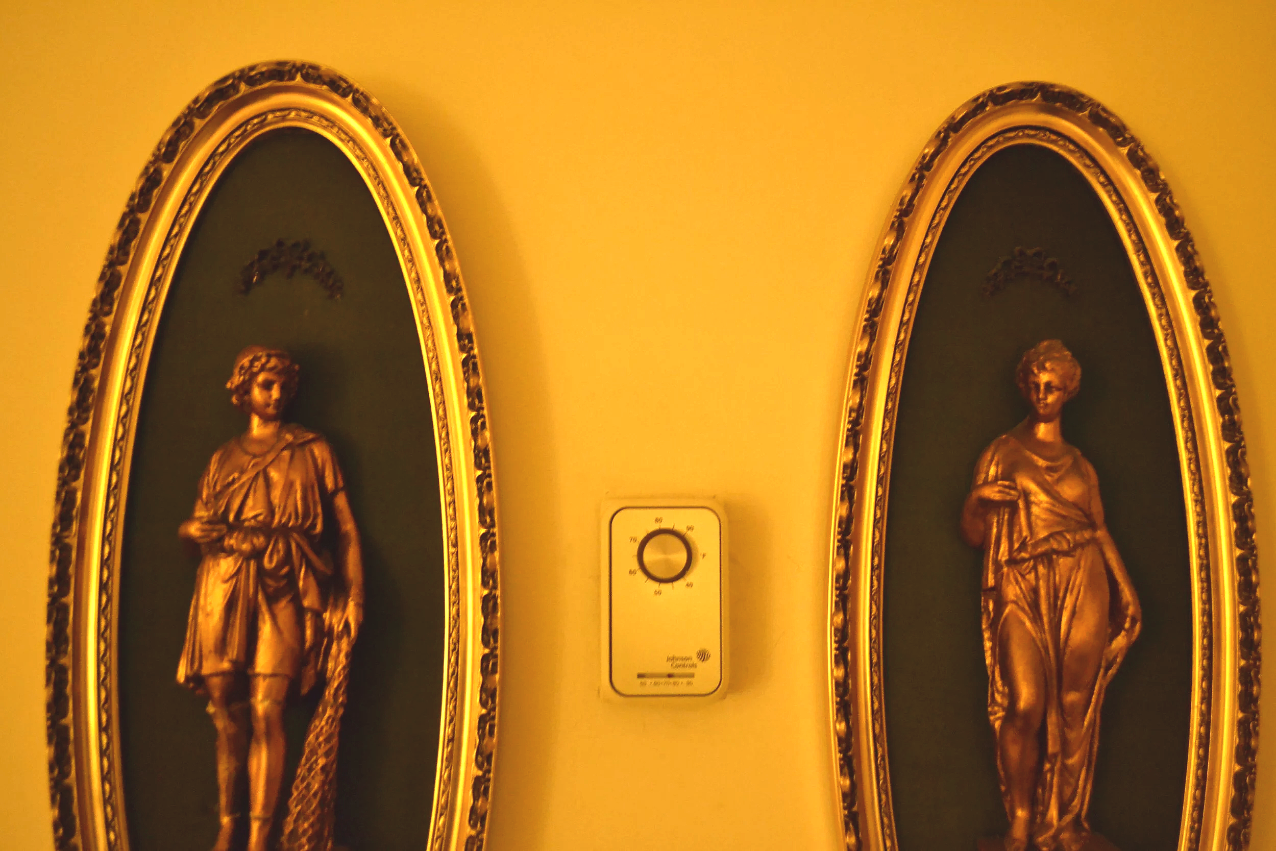 Close-up of two gold statues of women in ornate frames on a yellow wall, with a thermostat in between.