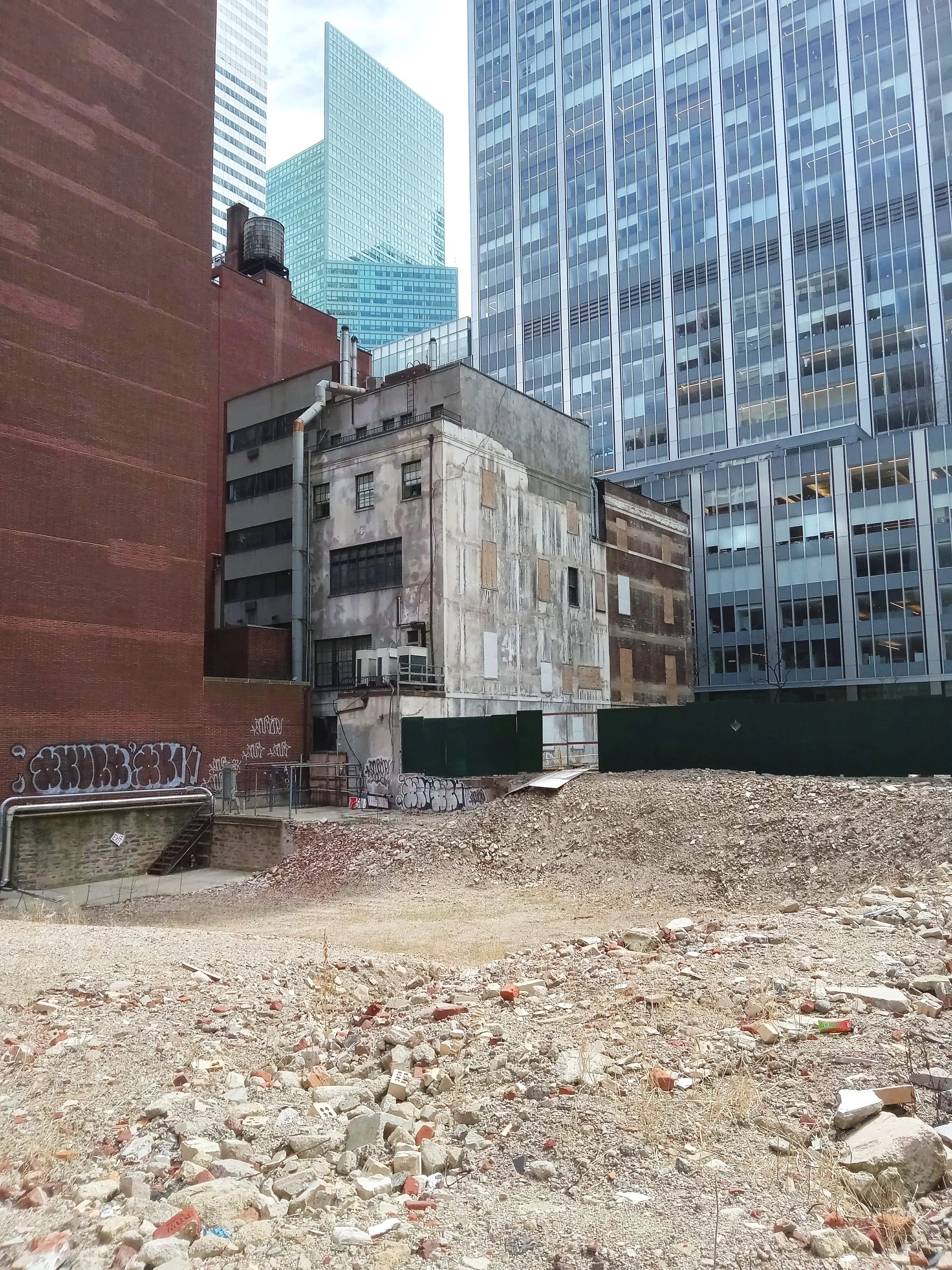 A city scene showing an empty lot with rubble and debris in the foreground, surrounded by older and modern high-rise buildings.