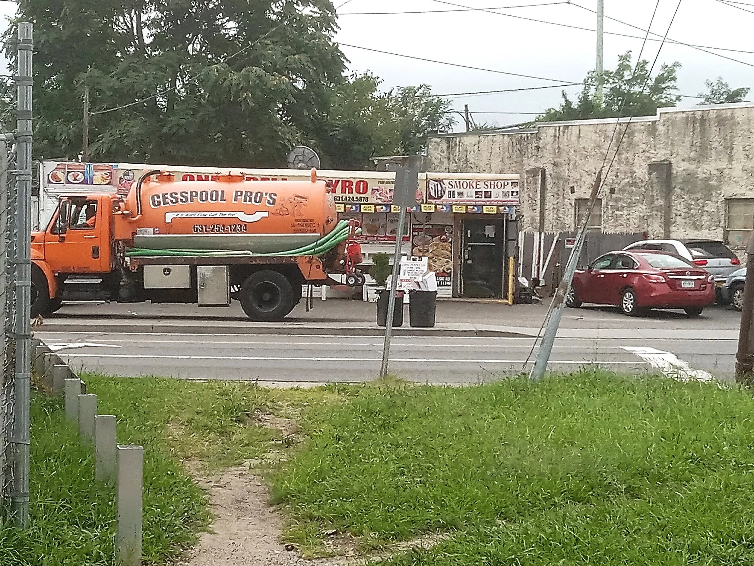 A street scene showing an orange truck with the label "Cesspool Pros" parked on the side of the road in front of a gyro shop with cars parked along the street, trees, and utility poles with power lines overhead.