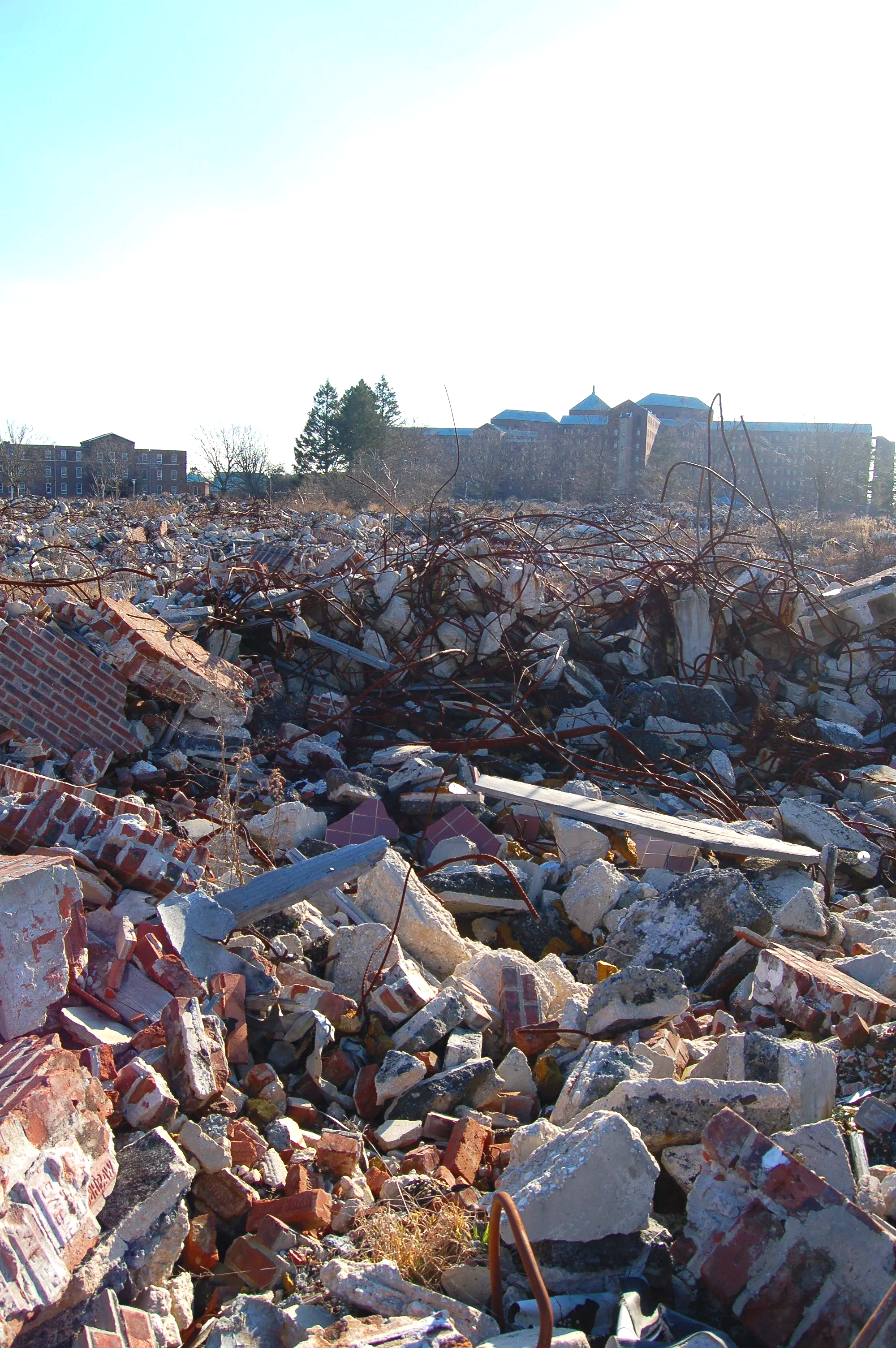 A large area filled with debris and rubble, including broken bricks, concrete, and twisted metal, set against a backdrop of buildings and trees under a bright sky.