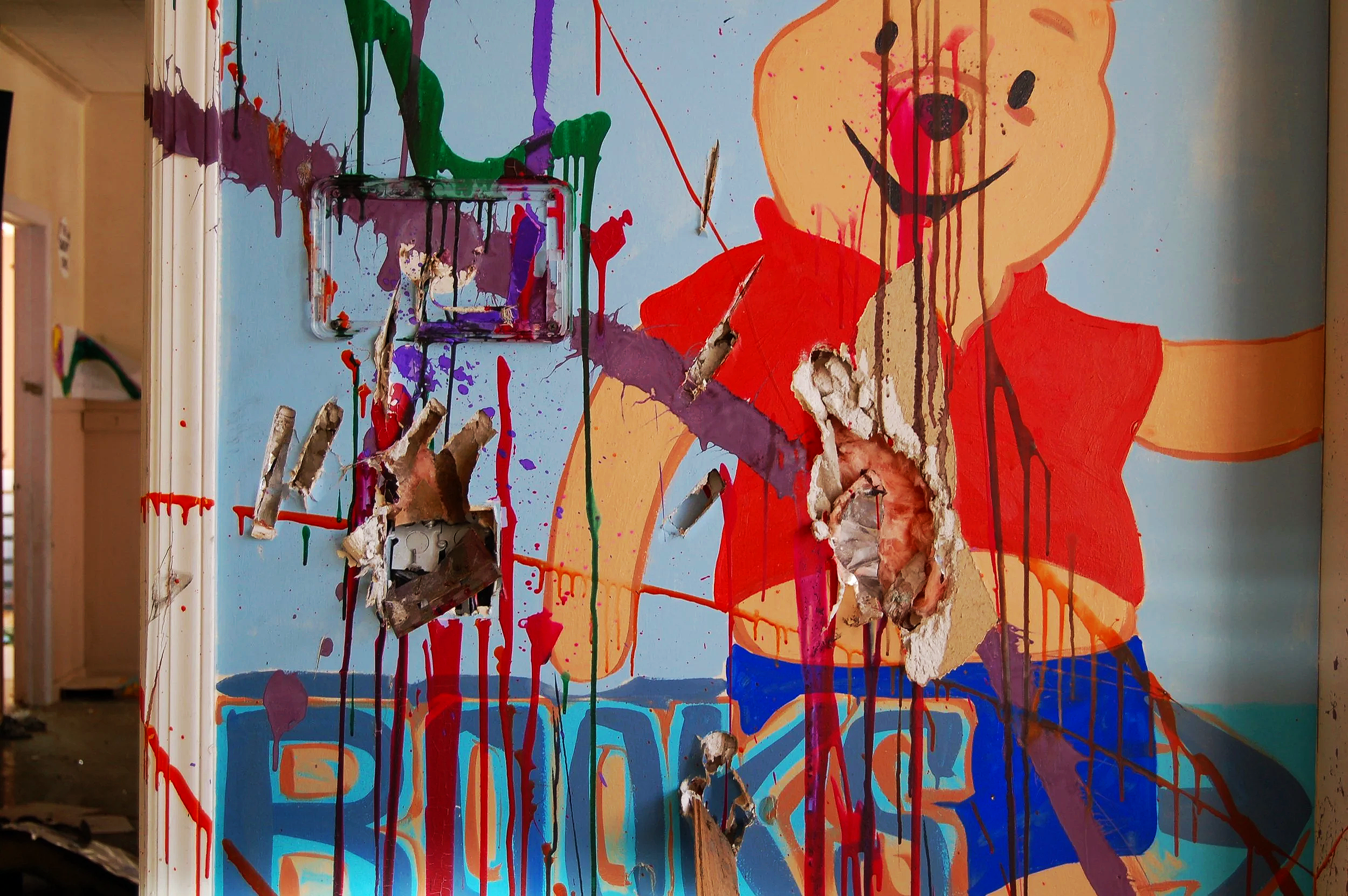 A colorful, abstract mural featuring a smiling cartoon bear with a red shirt on a blue background, partially destroyed with holes and torn paint revealing damaged drywall behind.