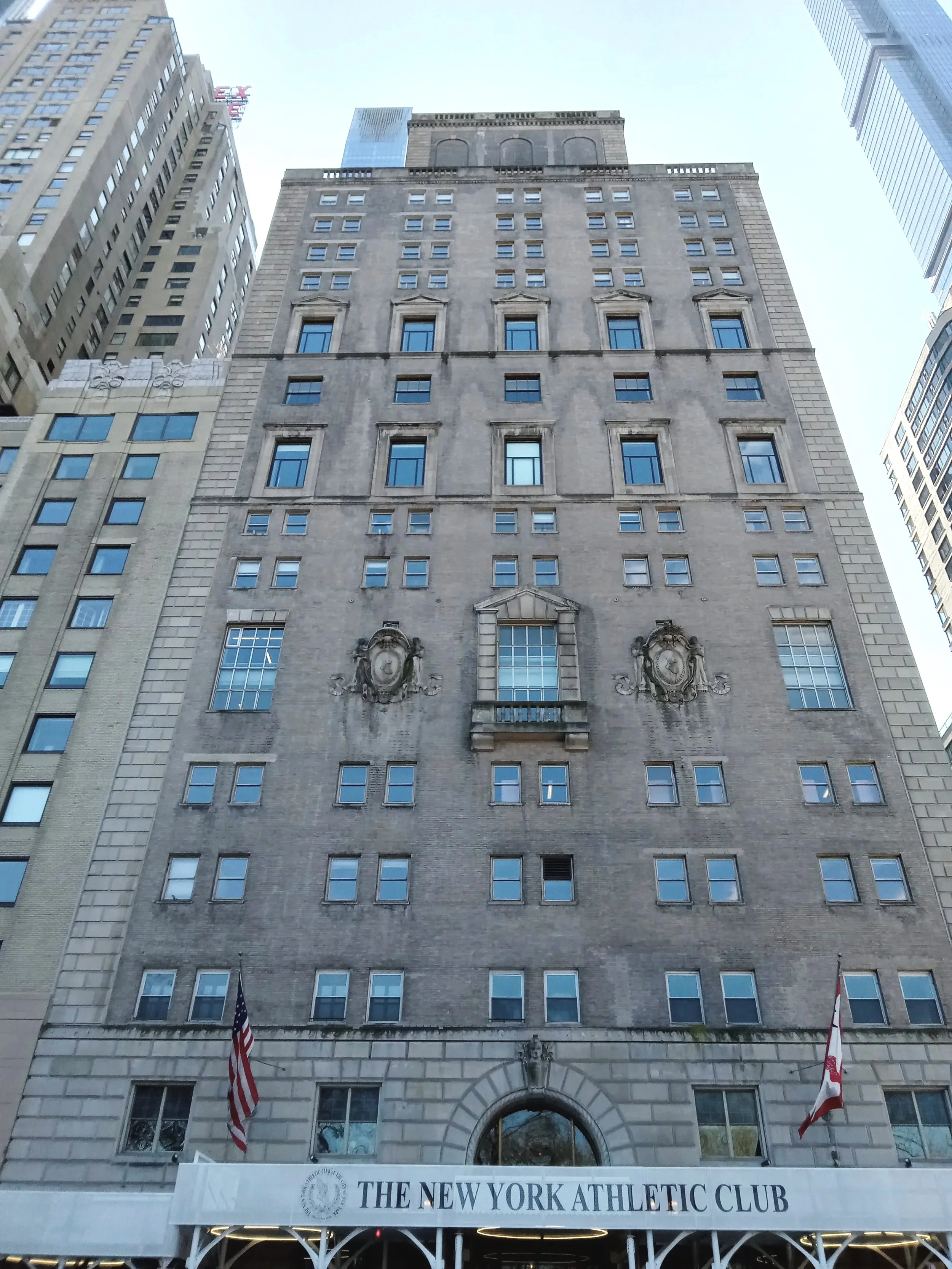 The photo shows the facade of the New York Athletic Club building, a tall stone structure with several windows. The entrance has a white awning with the words 'The New York Athletic Club' and flags on either side.
