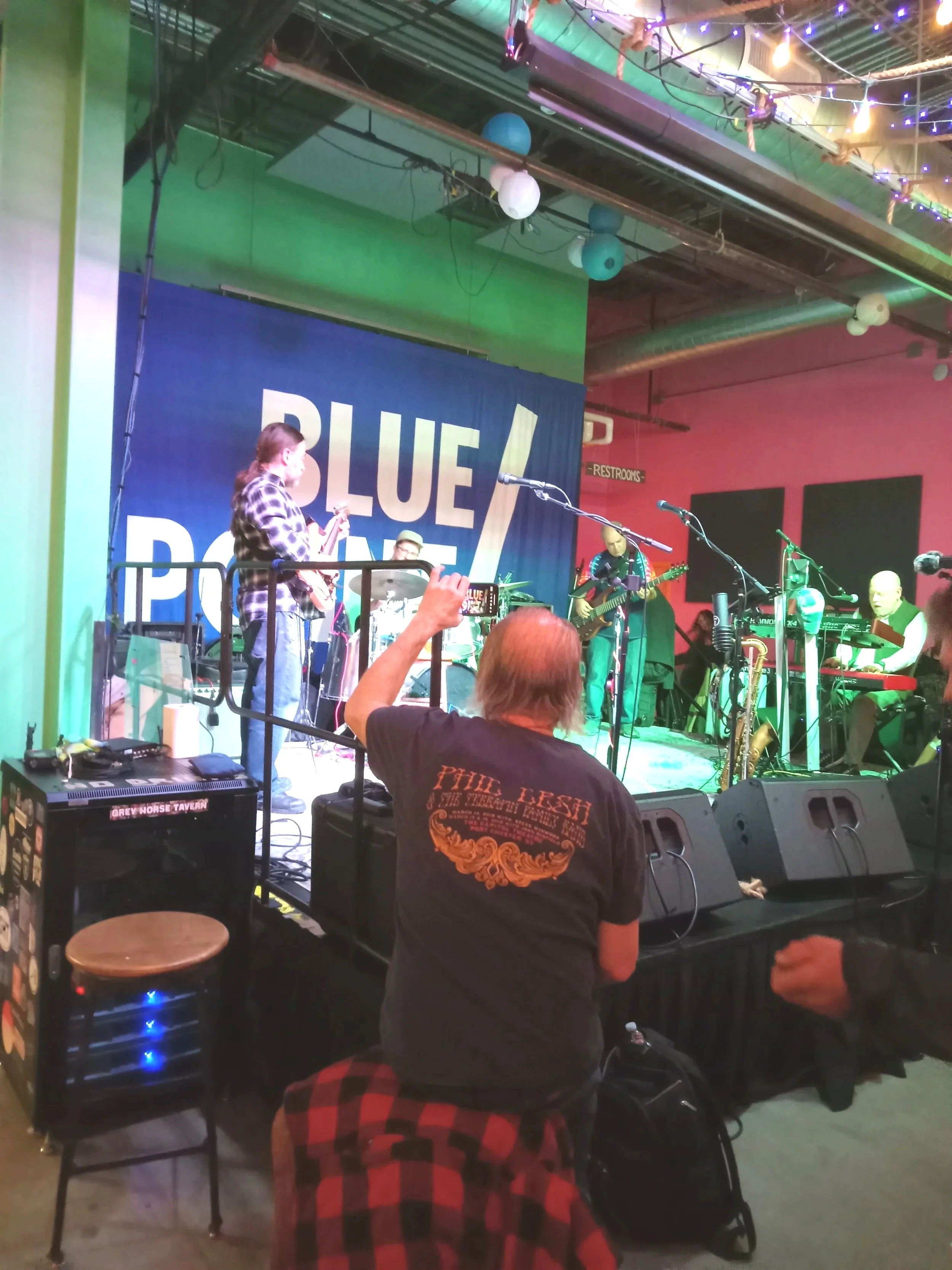 People playing musical instruments on stage at a live performance event. There are colorful string lights hanging above and a large blue banner with the word 'BLUE' visible behind the band. An old man sits and enjoys the music.
