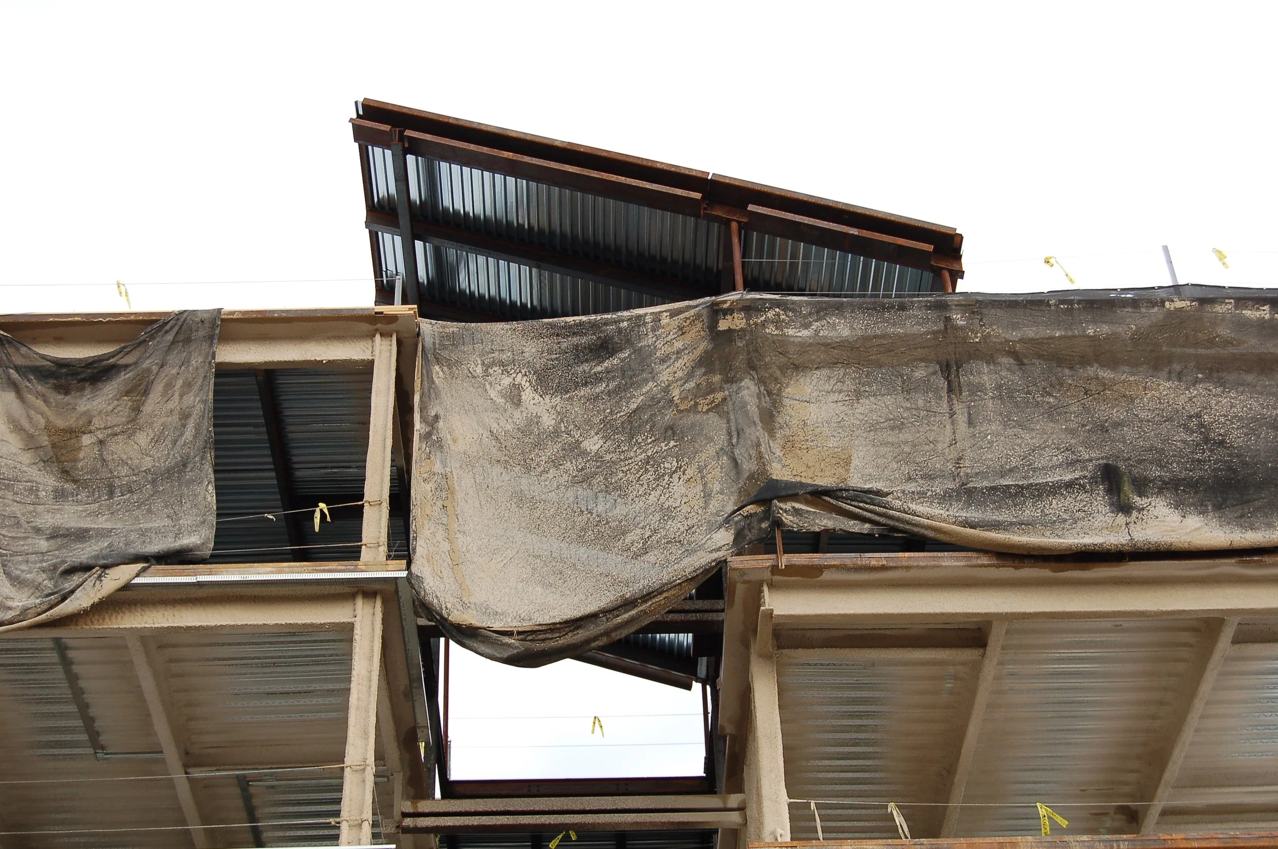 A building under construction with exposed concrete and steel beams, covered partially by black construction fabric, and a roof made of metal panels.