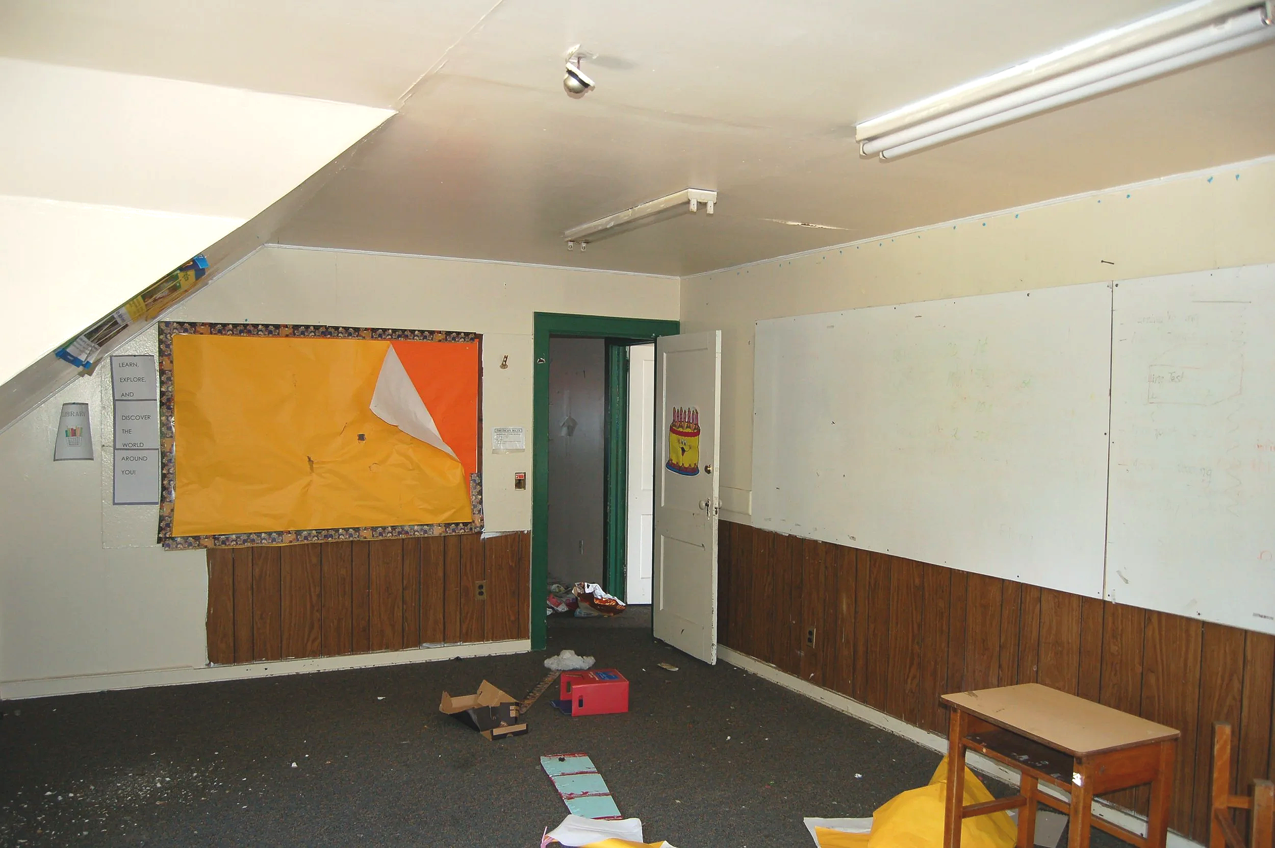 A classroom with a whiteboard on the wall, a bulletin board with orange paper, and scattered papers on a black carpeted floor. The room has wood paneling at the bottom of the walls and white paint above. There is an open door with visible clutter inside, and a small wooden table near the wall.
