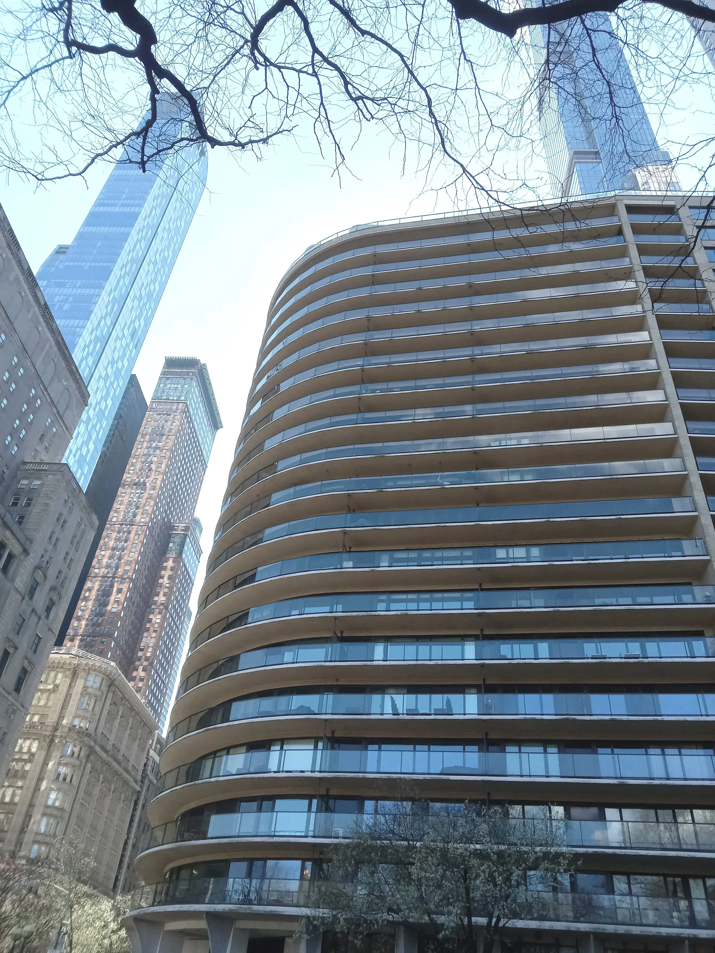 Tall modern skyscrapers with glass facades and curved balconies, city street view with trees and partial buildings visible.