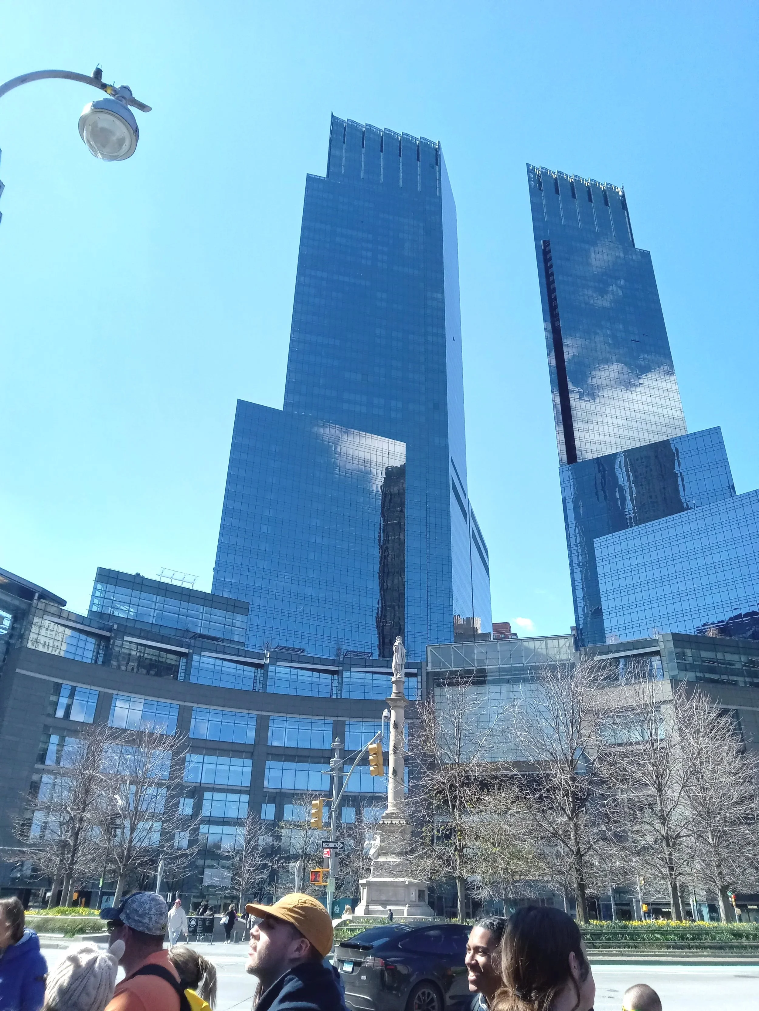 People walking on city street with tall modern glass skyscrapers in background, blue sky, traffic lights, and trees.