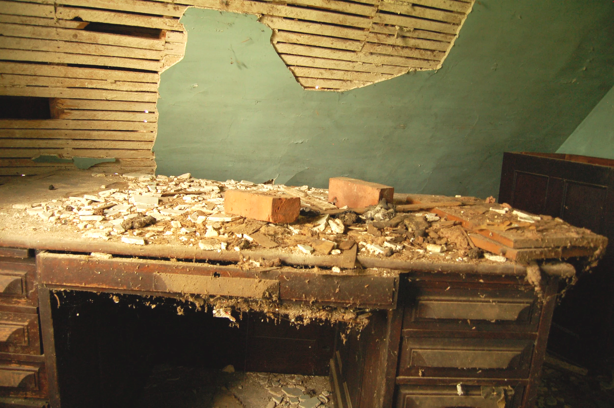 A ceiling with peeling paint and exposed wooden slats, debris scattered on a surface below, and a partial view of furniture in the corner.