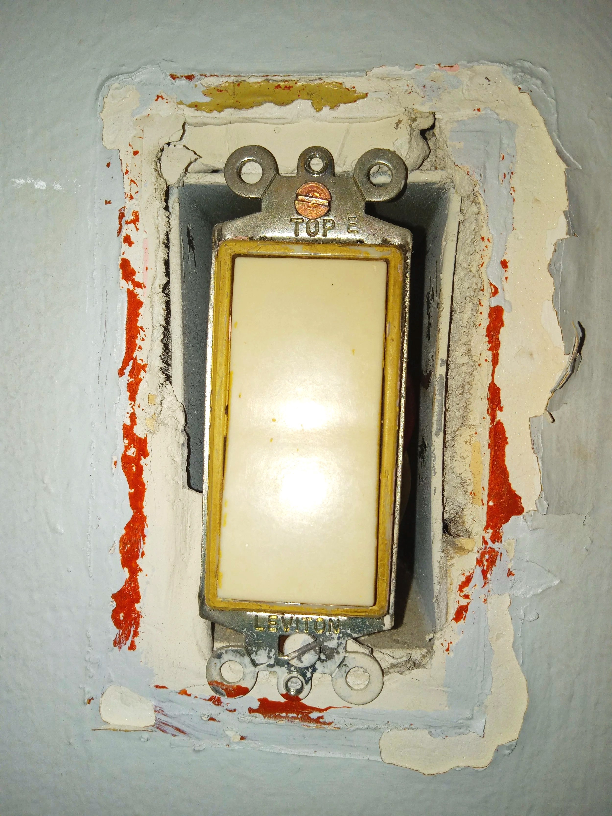 Close-up of an old electrical outlet box with a mounted blank switch plate on a wall with peeling paint and damaged plaster.