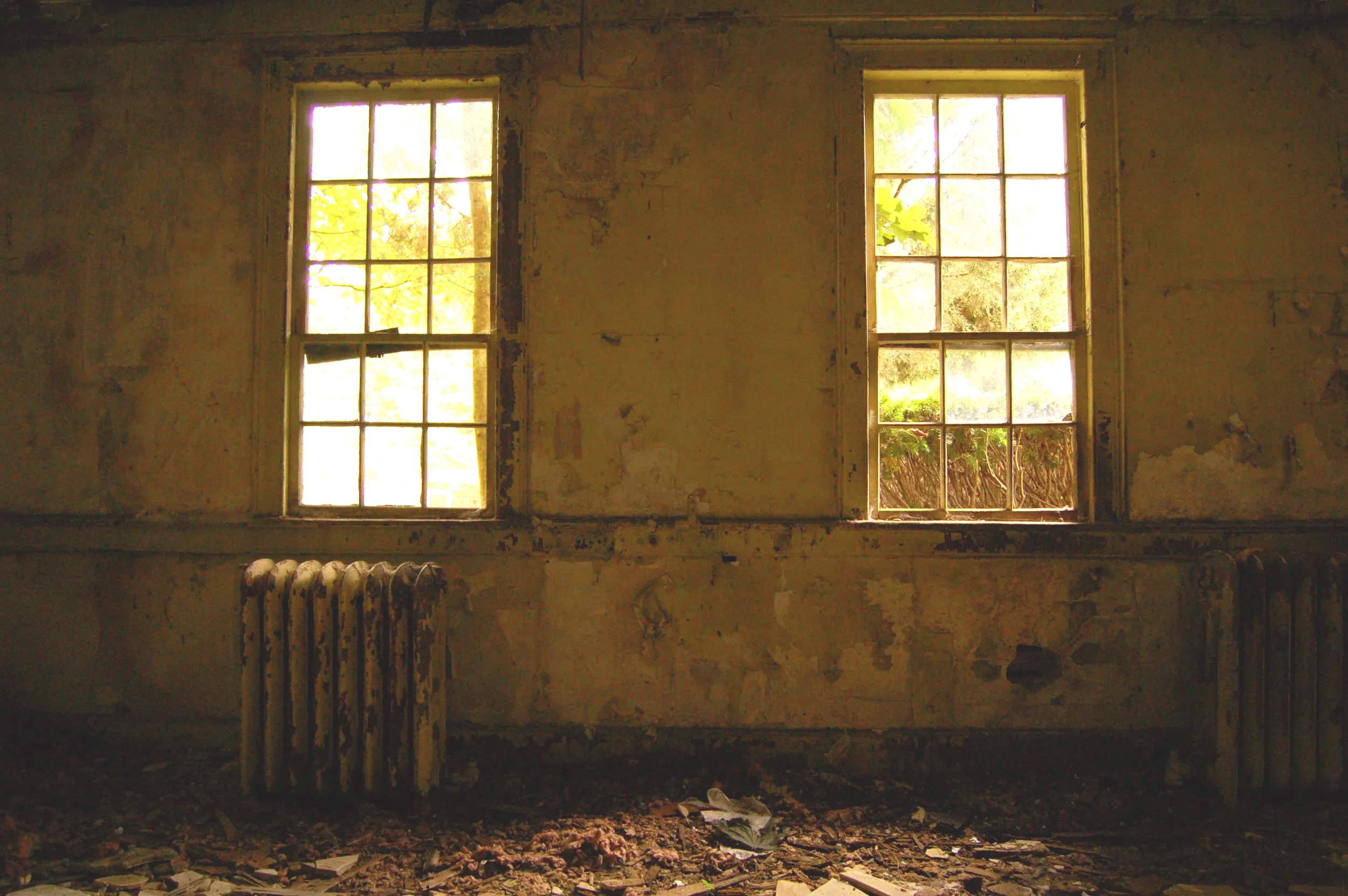 Interior view of an abandoned building with two old windows letting in natural light, a rusty radiator beneath the left window, and debris on the dusty floor.