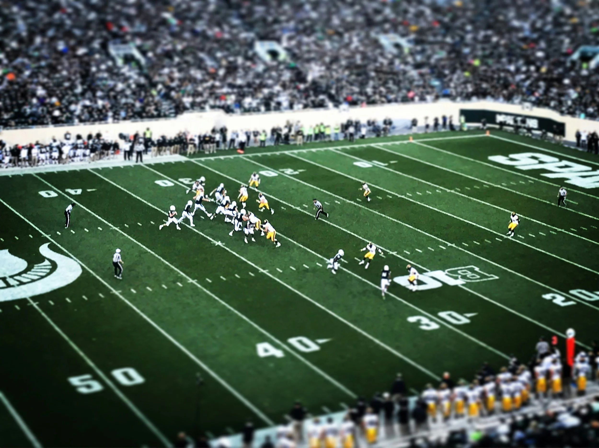 Football game on a green field with players and officials, packed stadium with spectators.