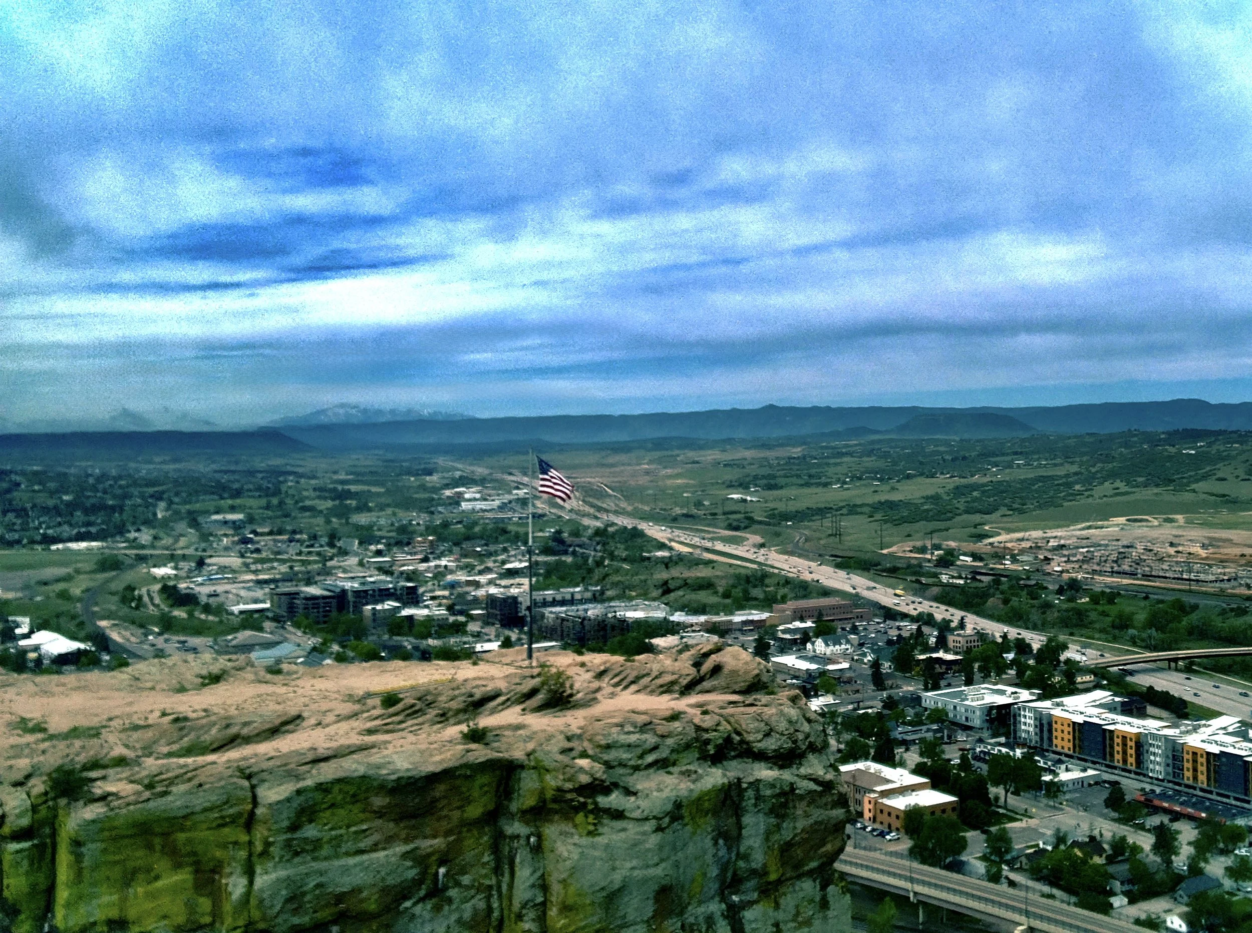 An American flag flying over a small cityscape with buildings, roads, and a distant mountain range under a cloudy sky.