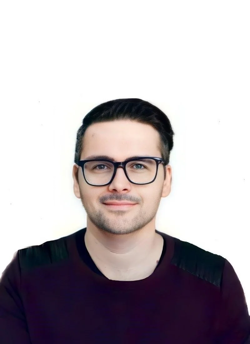 Portrait of a young man with dark hair, glasses, and a slight smile, wearing a black shirt with leather patches on the shoulders against a plain white background.