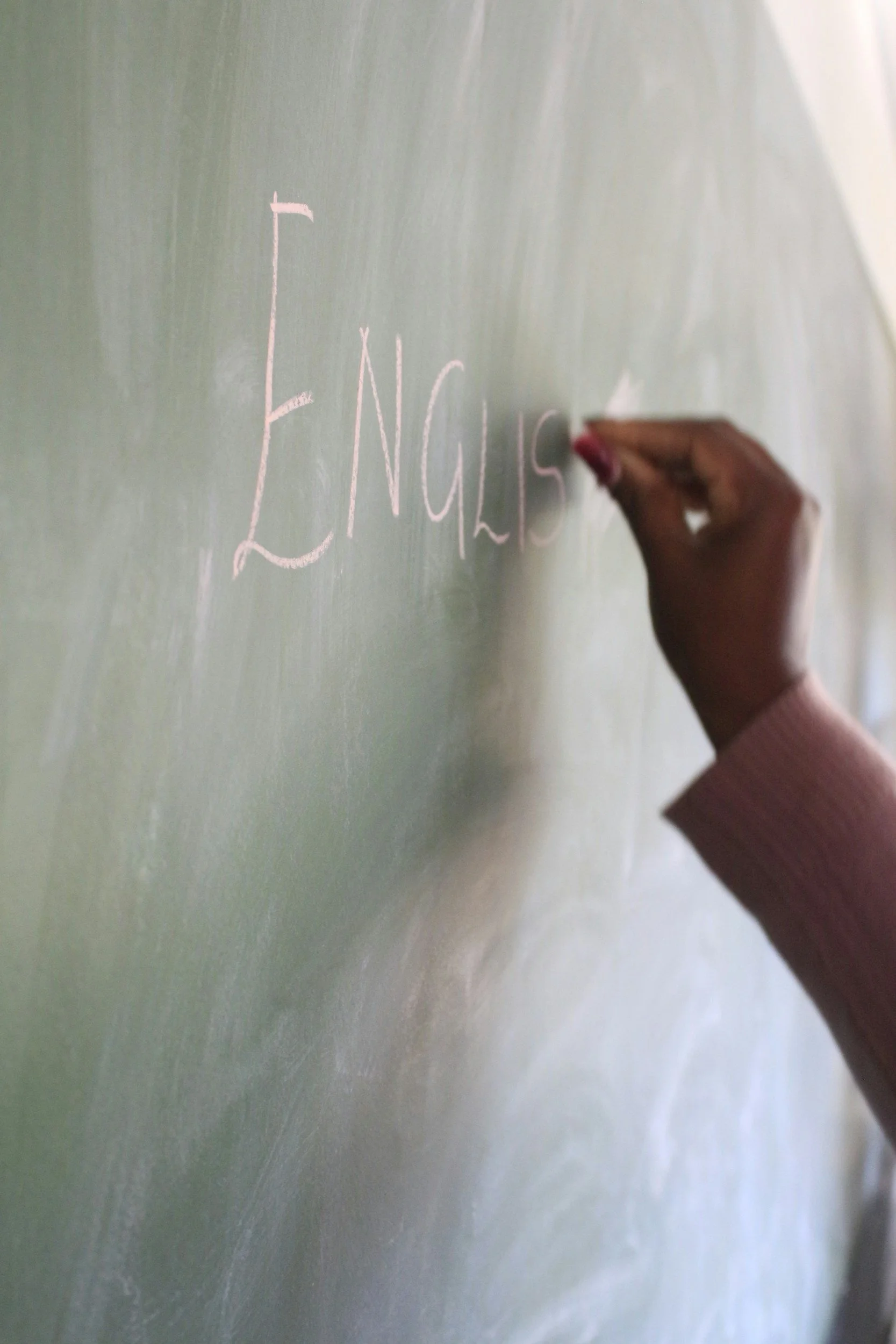 Close-up of a hand writing on a green chalkboard with pink chalk. The hand appears to be writing the word 'English'.