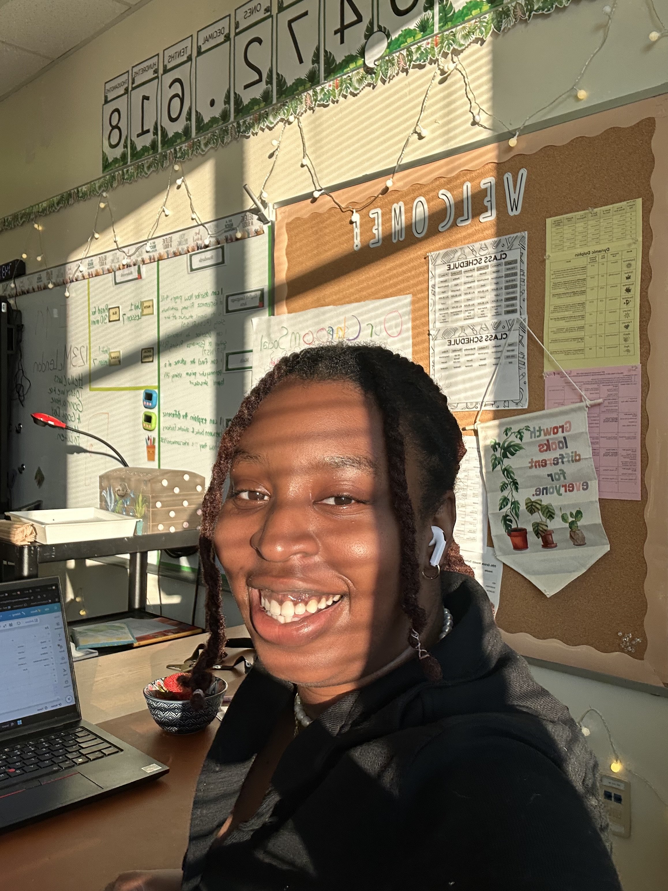A person smiling in an office with a bulletin board, string lights, and a computer on the desk.