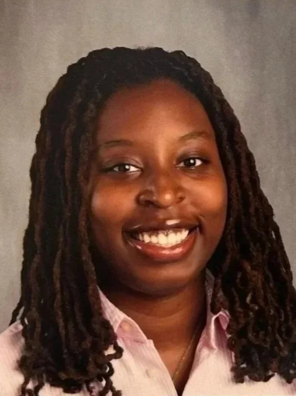 A young woman with dark skin and long twisted dreadlocks smiling in front of a grey background.