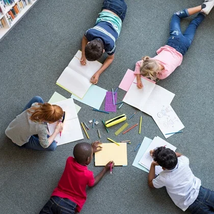 Five children lying on the floor, drawing and coloring with markers, surrounded by opened notebooks and drawing supplies.