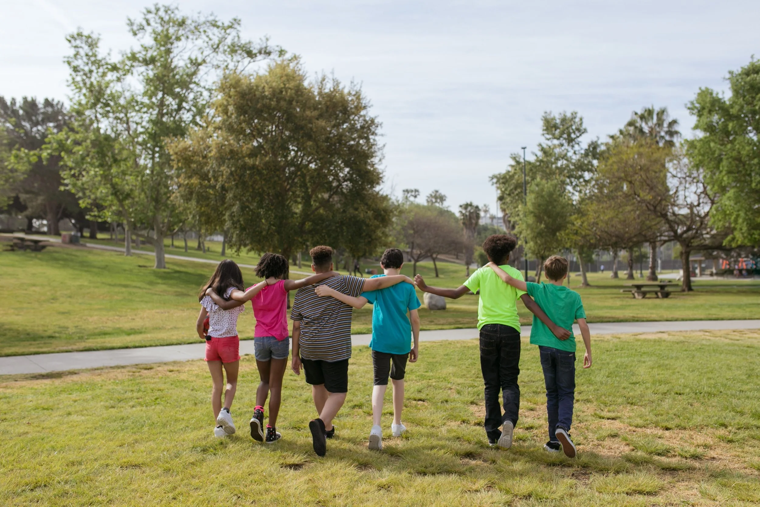 A group of children walking arm-in-arm through a park on a cloudy day with trees and grass in the background.