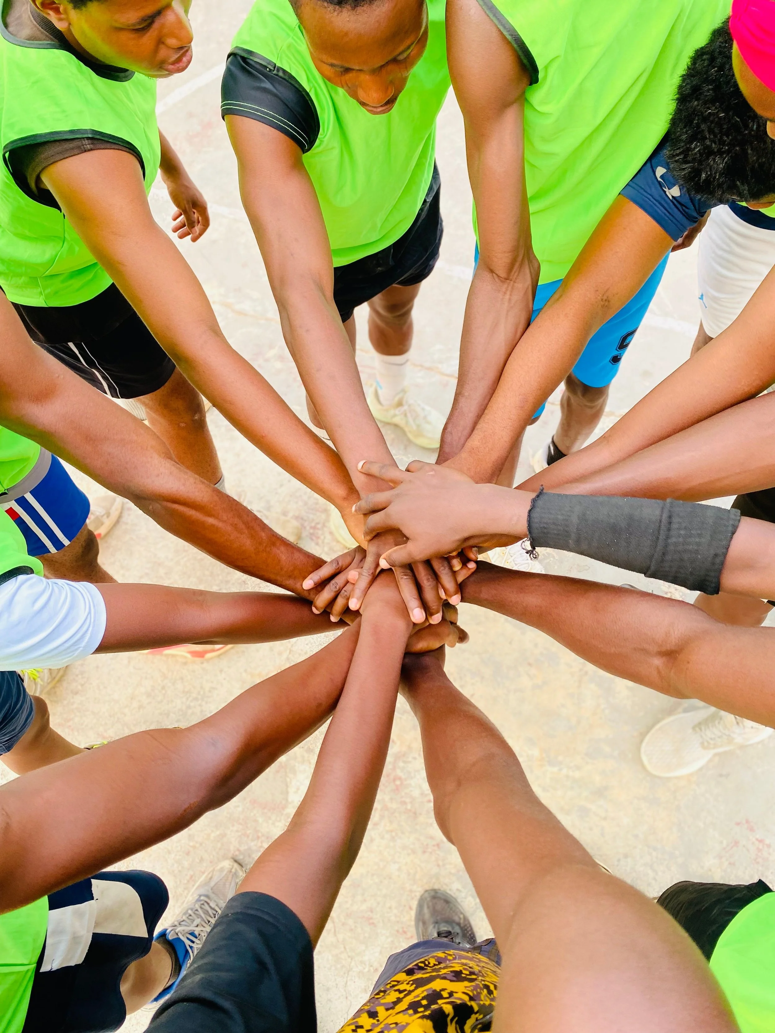 Group of people with varying skin tones in neon green jerseys, joining hands in the center for a team gesture, on a sports court.
