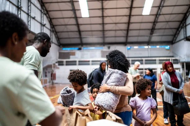 People, including children and adults, gather inside a gymnasium, carrying bags and personal belongings.